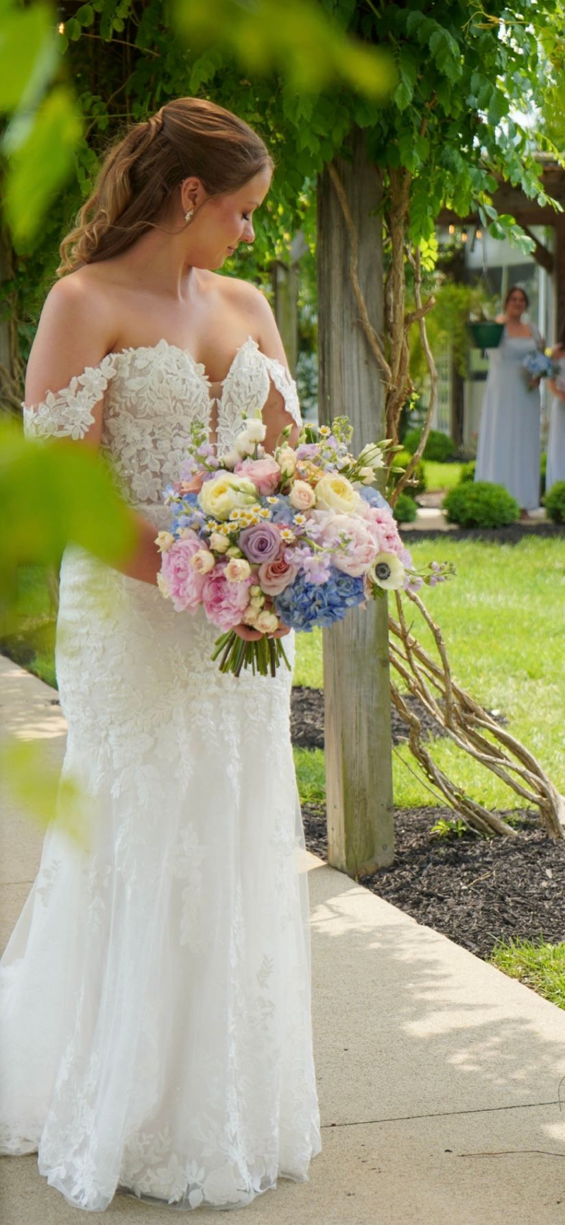 Bride in off-shoulder lace wedding gown holding pastel floral bouquet outdoors under green arbor.