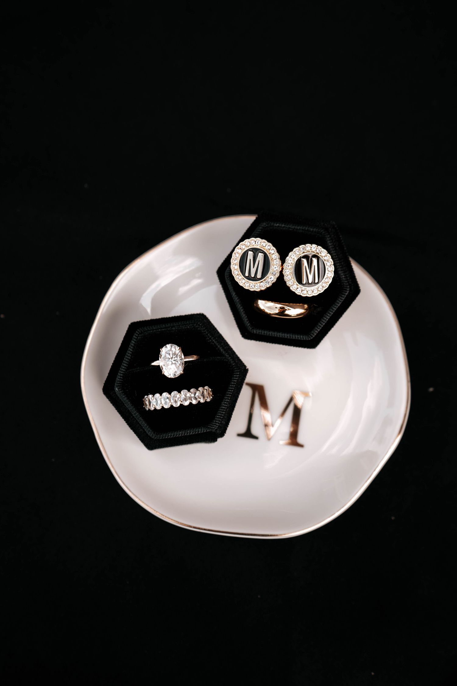 Wedding rings and monogram M cufflinks displayed in black velvet boxes on white ceramic plate.
