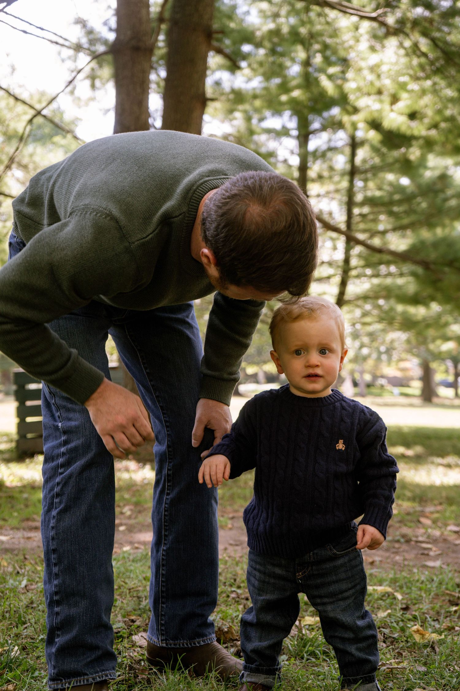 Father bending down to interact with his curious toddler son in a green outdoor park setting.