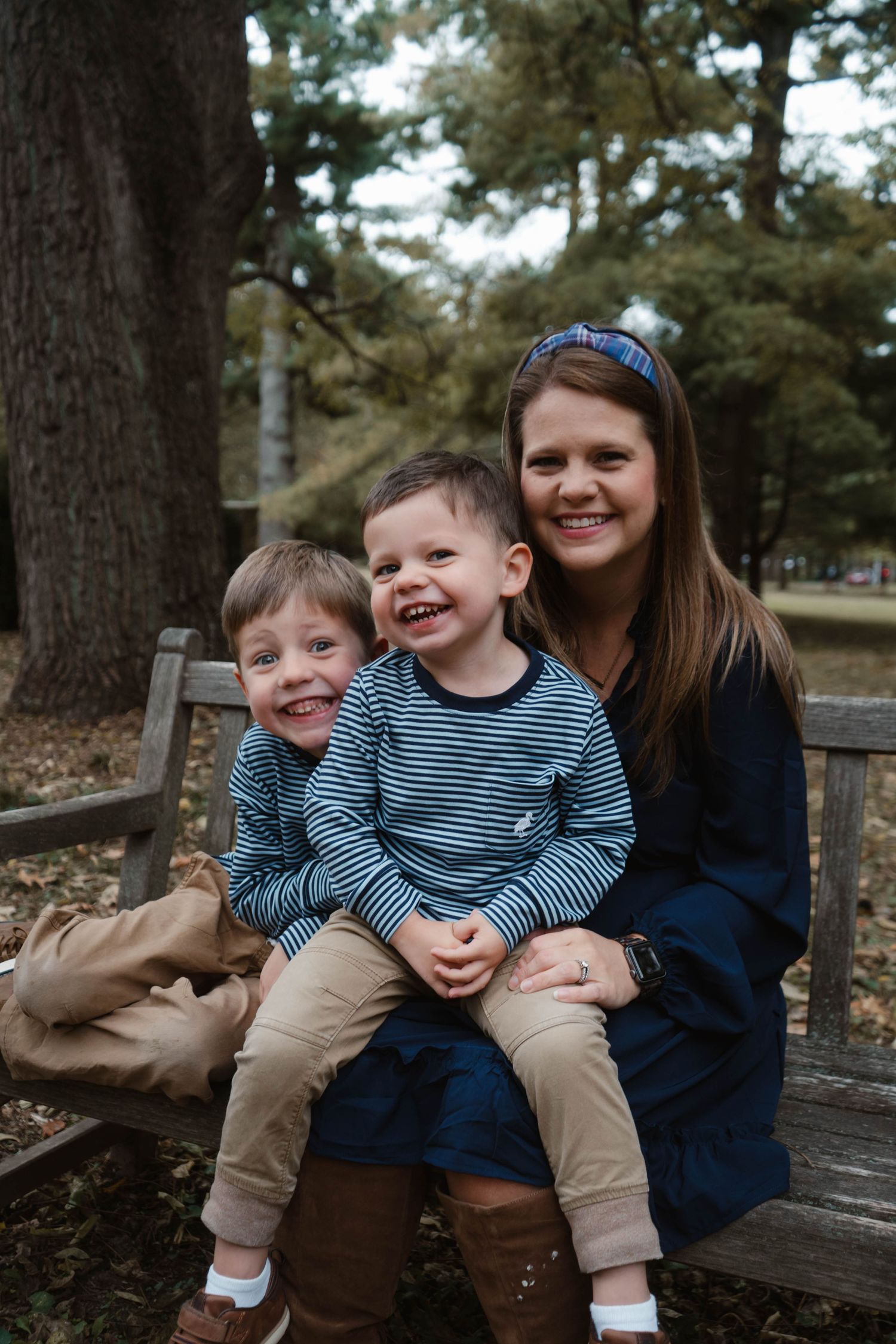 A smiling woman sits on a park bench with two young boys in matching striped shirts and khaki pants outdoors.
