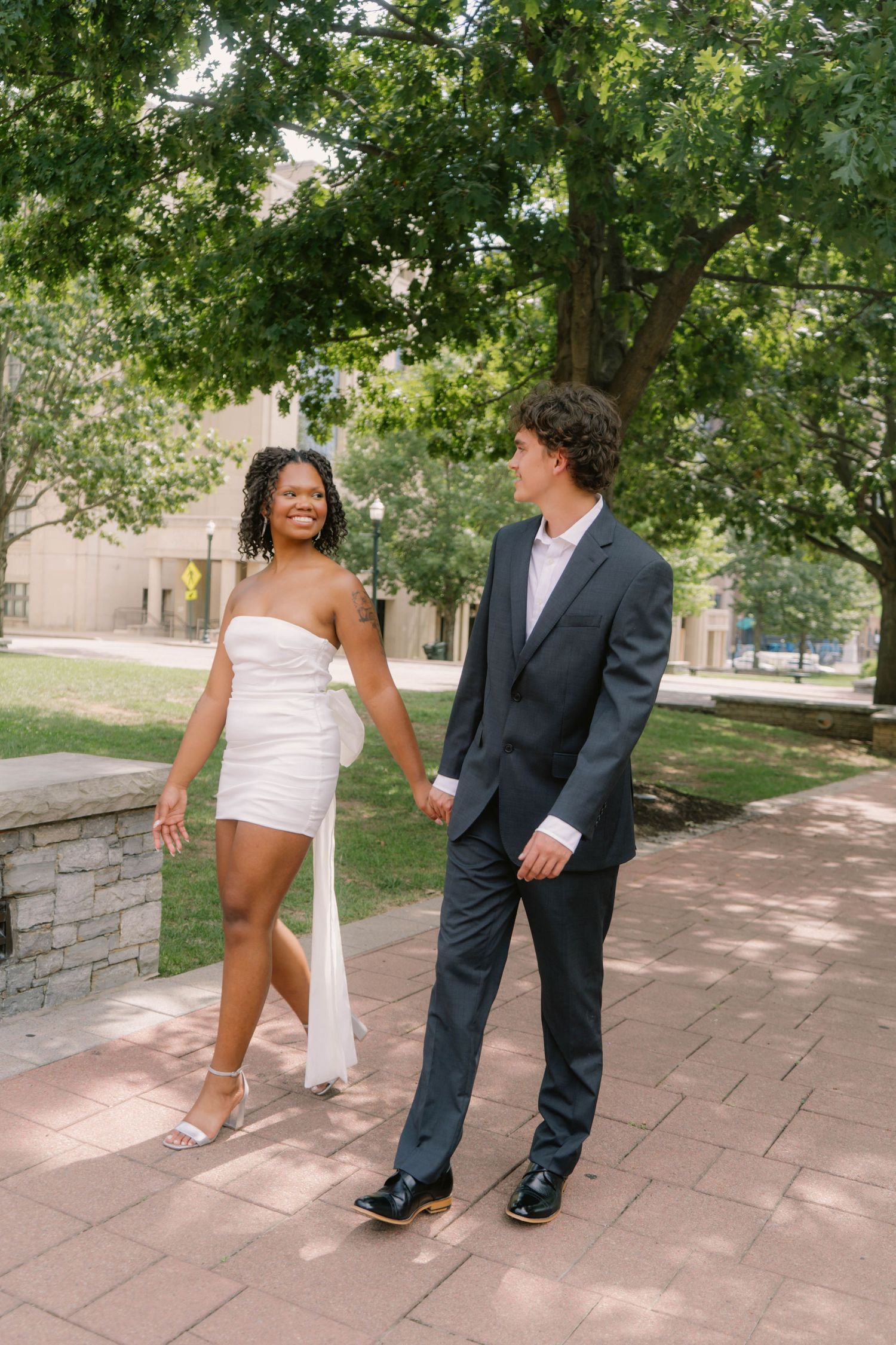 Joyful bride in strapless white mini dress leads groom by hand along stone pathway under green trees.