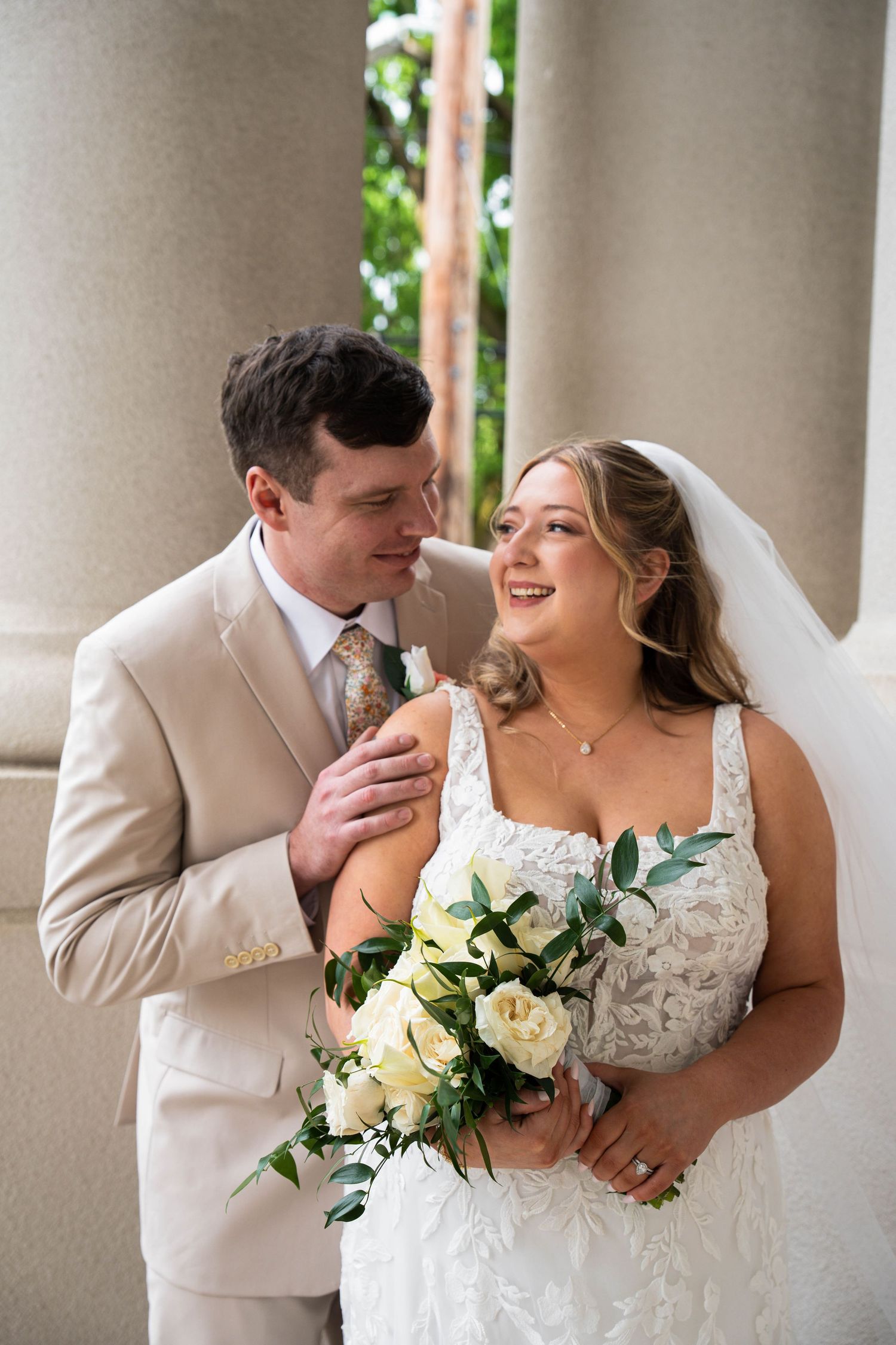 Bride and groom laugh together outdoors, she holds white roses, he wears a tan suit with floral tie.