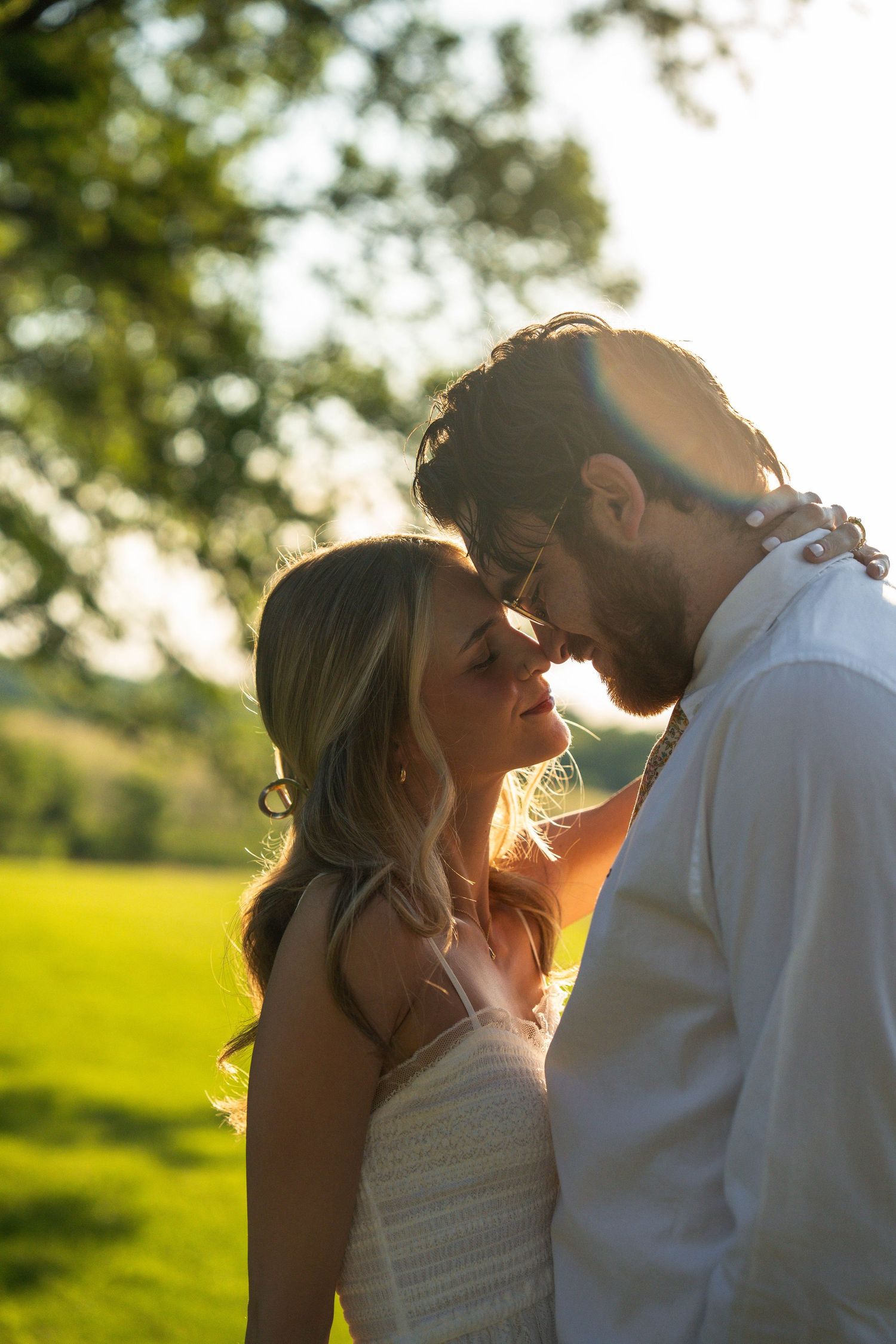 Bride and groom touch foreheads romantically in golden sunset light beneath a lush green tree.