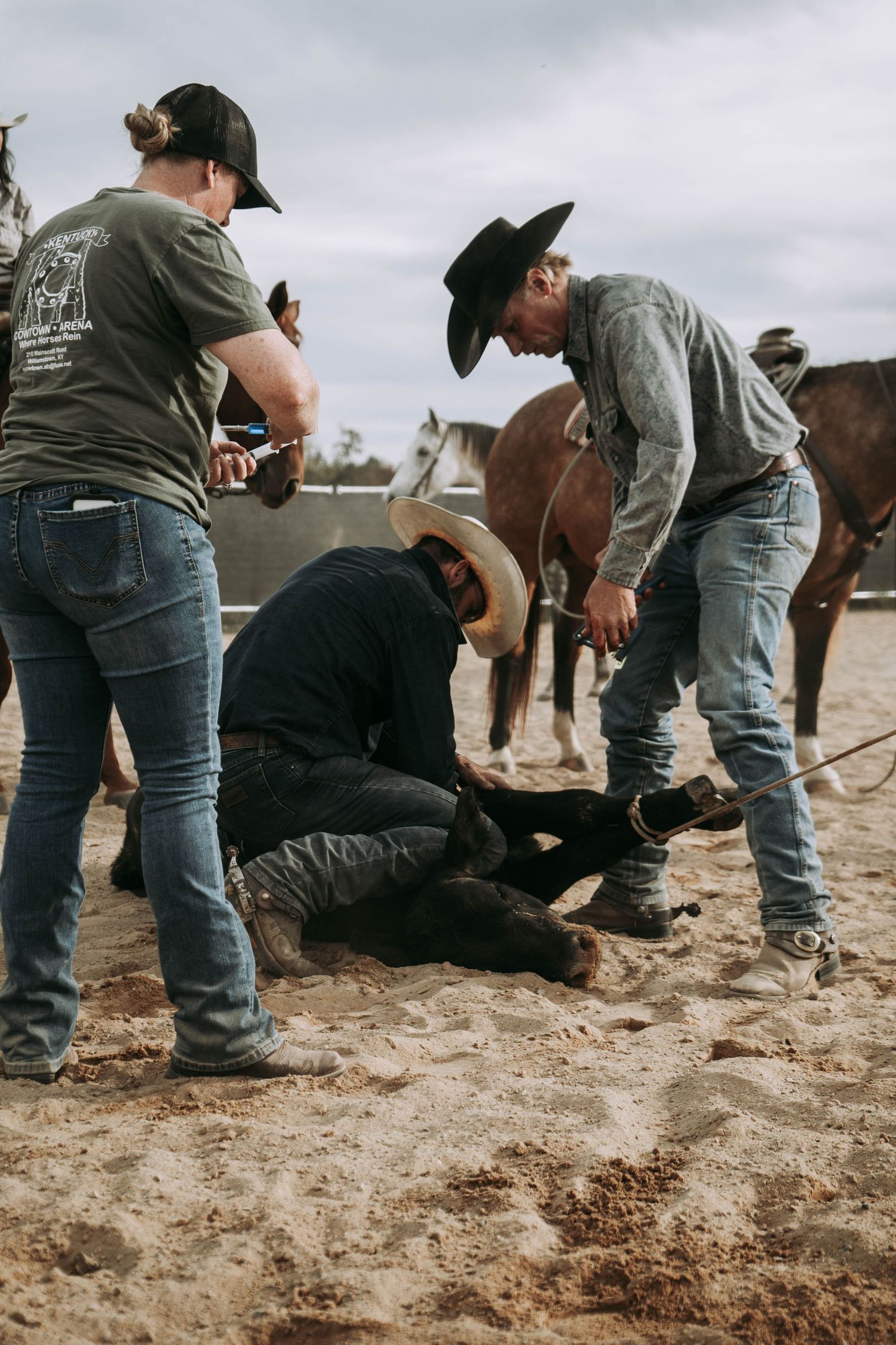 Cowboys working together to rope and handle a calf in a sandy rodeo arena with horses nearby.