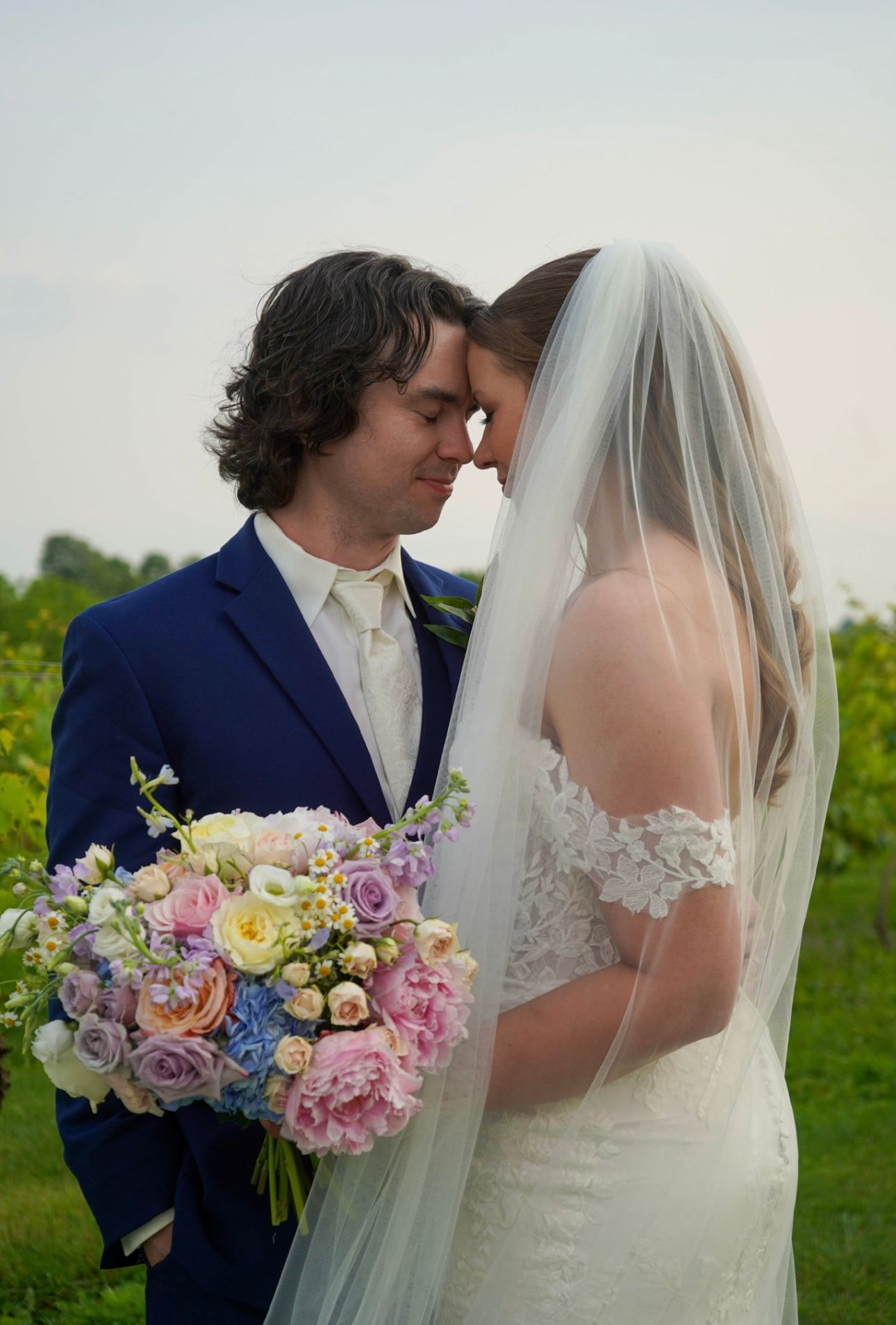 Romantic bride and groom touching foreheads in vineyard, bride wearing lace gown holding pastel spring bouquet.