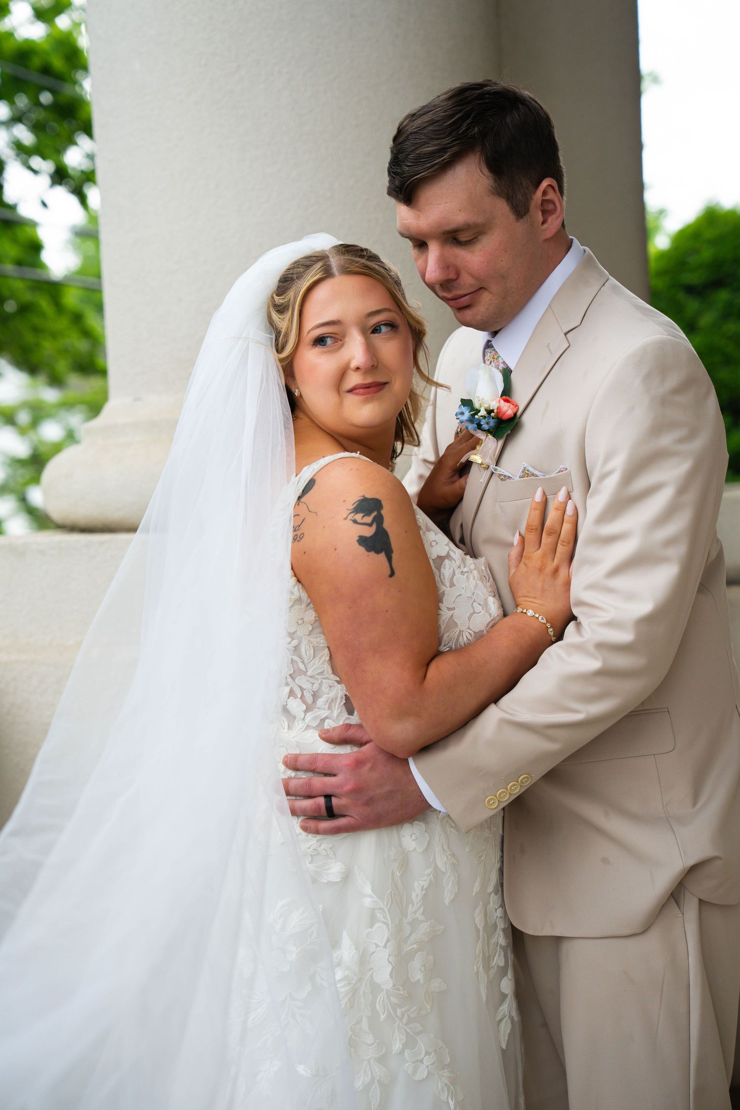 Bride and groom embracing by stone columns, bride wearing lace gown with floral arm tattoo.