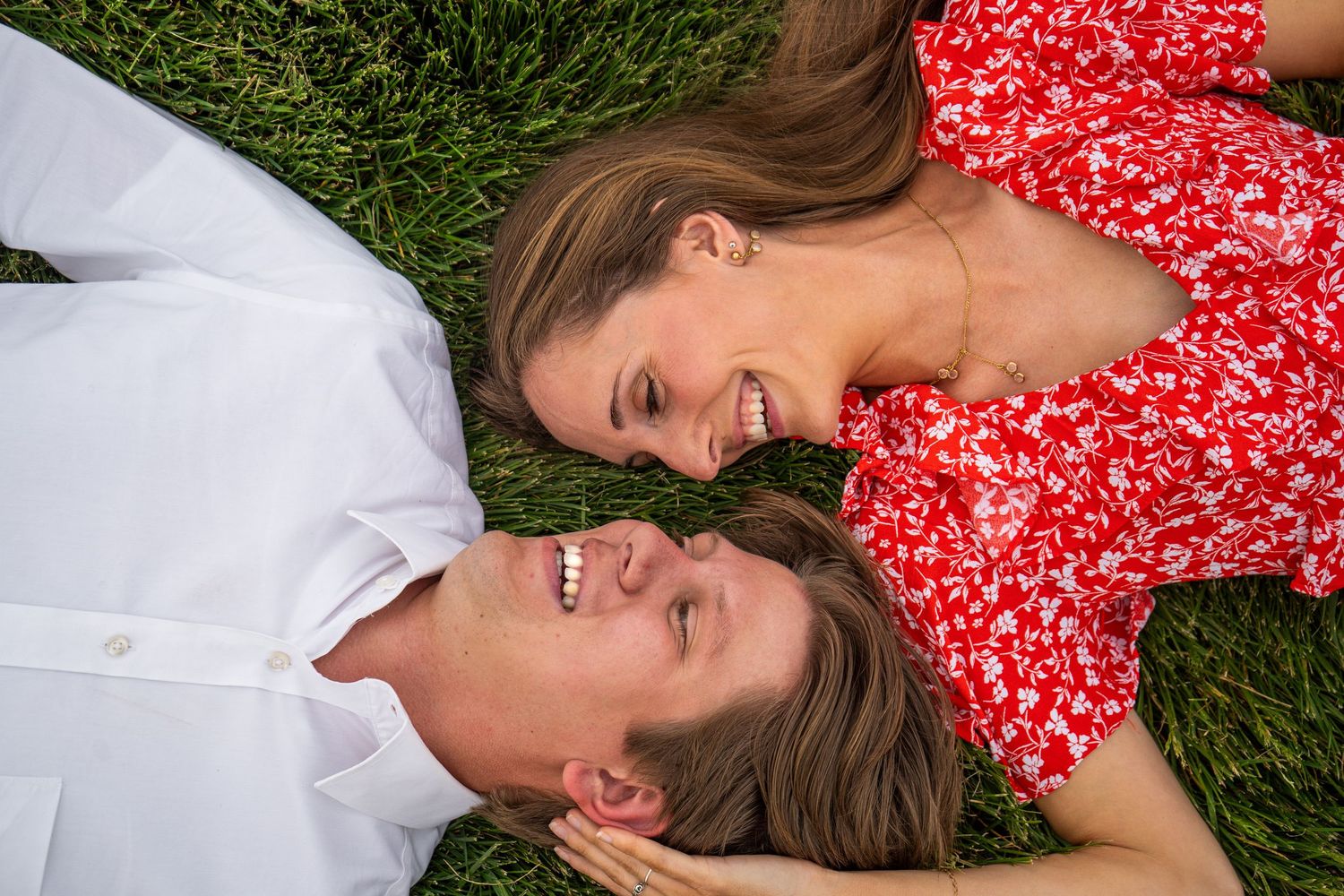 Couple lying on green grass smiling at each other, woman in red floral dress, man in white shirt.