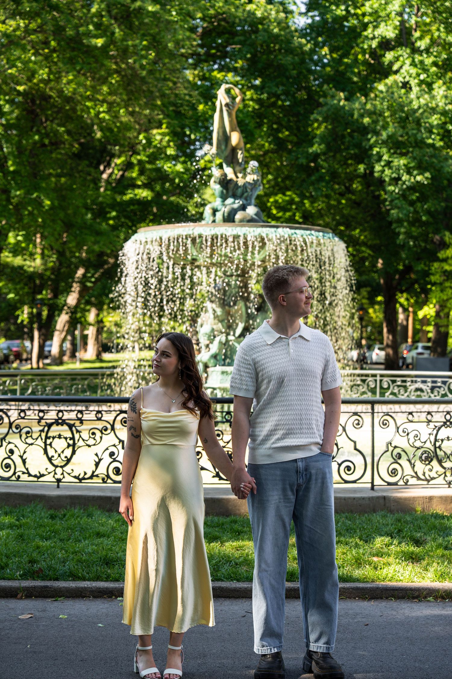 Engaged couple holding hands in front of an ornate fountain in a lush green park.