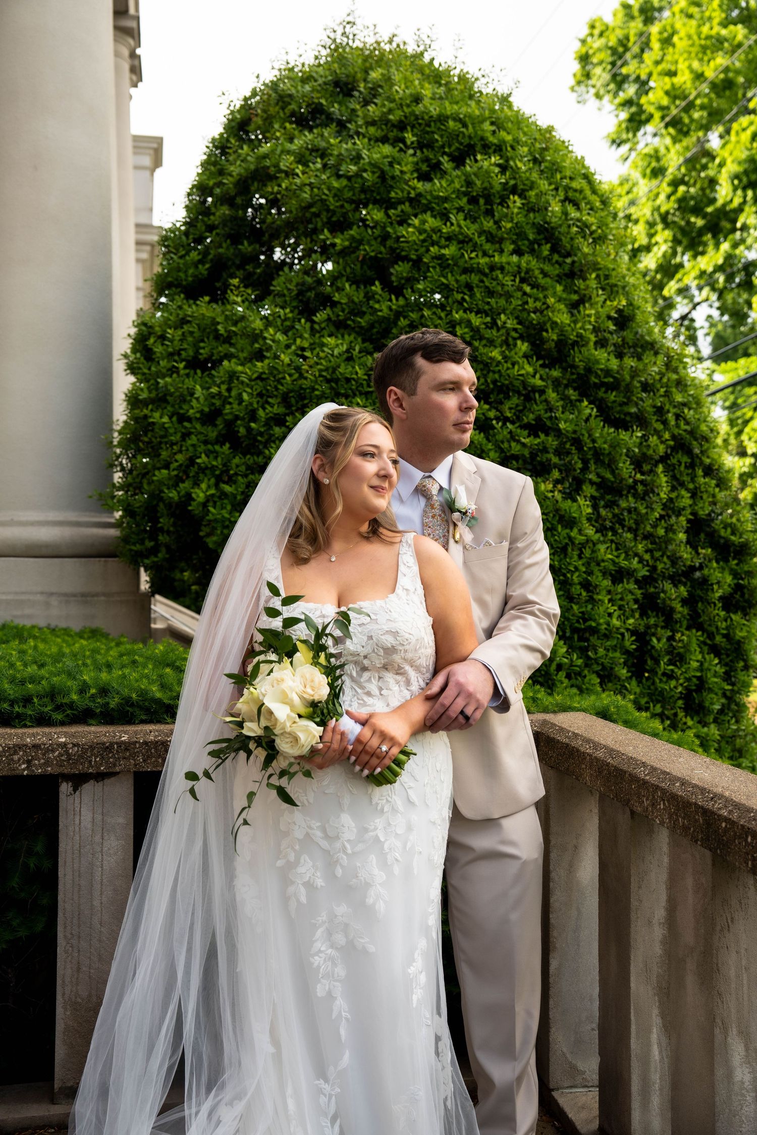 Elegant bride holding white bouquet and groom in tan suit pose together on a balcony surrounded by greenery.
