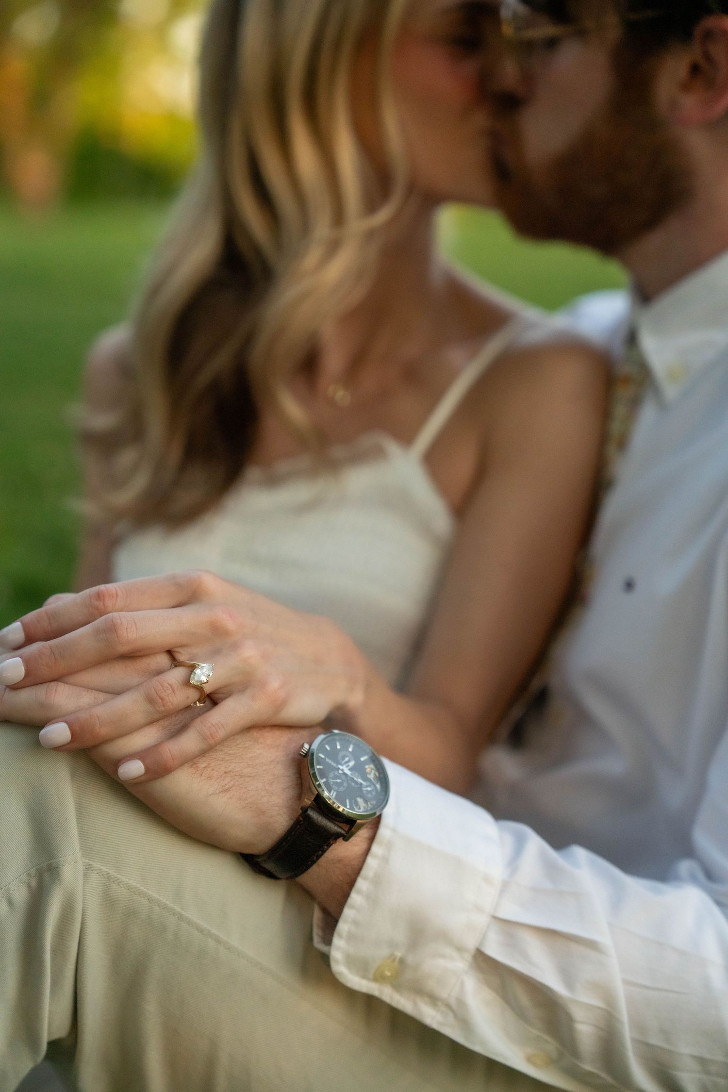 Close-up of engaged couple kissing, showcasing diamond engagement ring and mans leather strap watch.