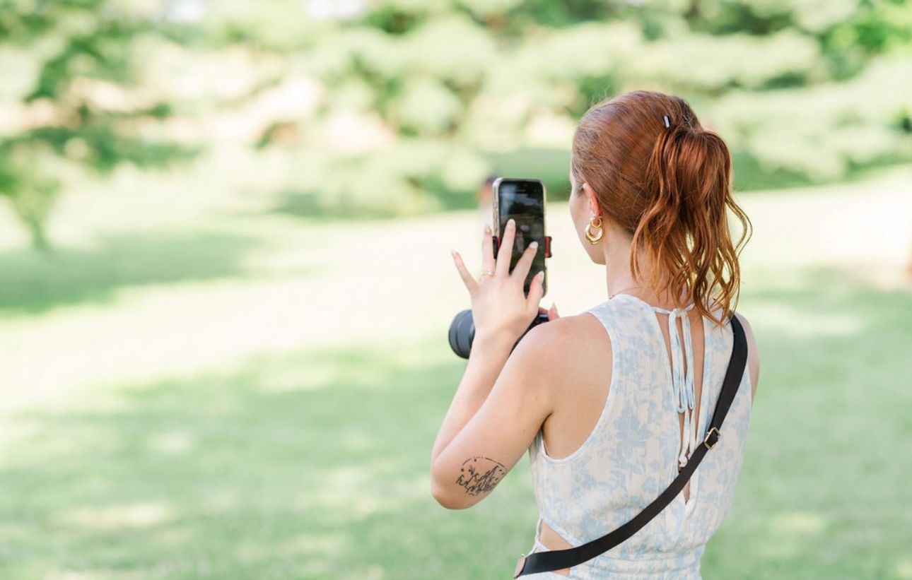 Red-haired woman with tattoo photographs outdoor scenery using smartphone in sunlit green park.