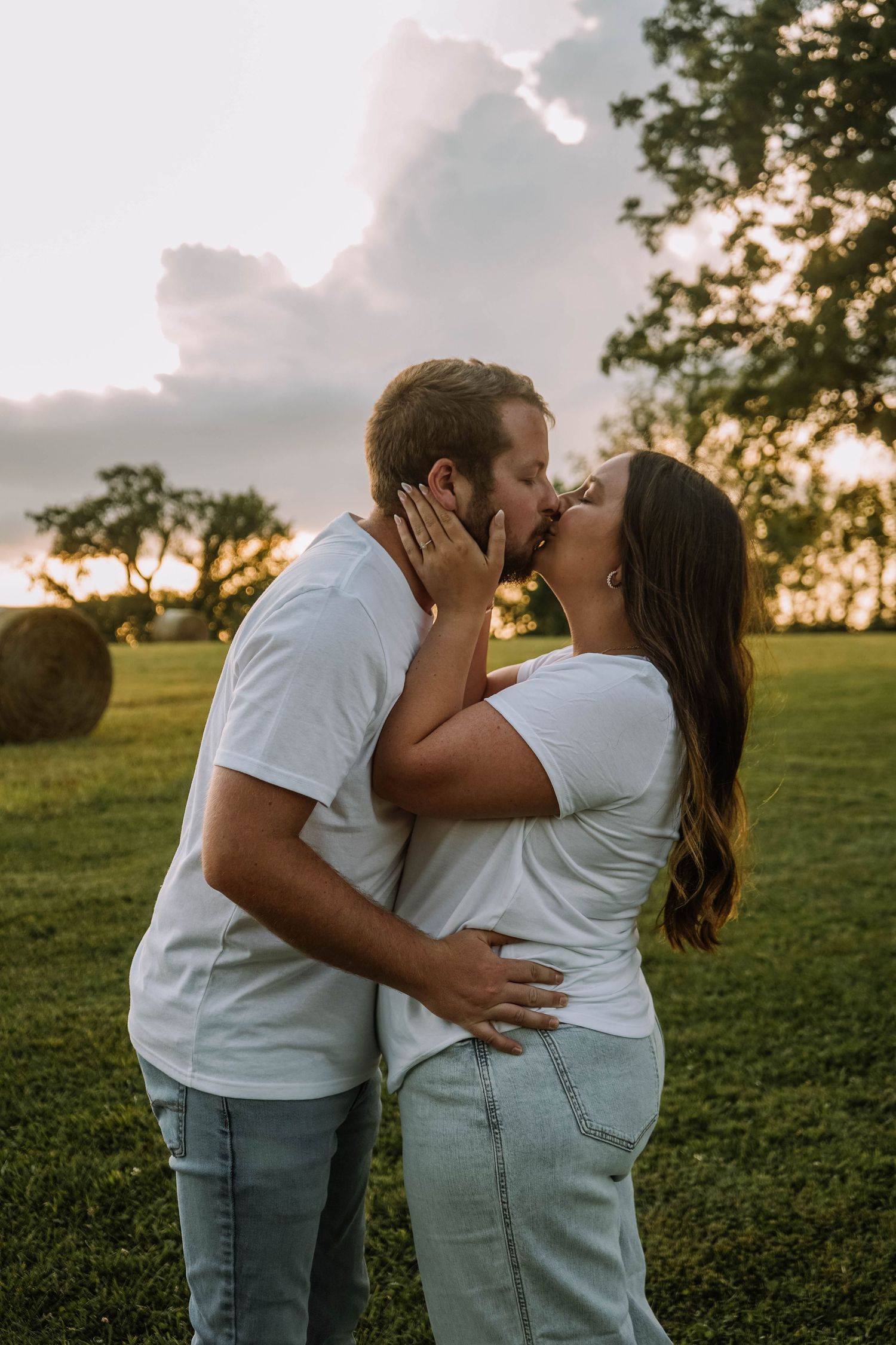 Couple in white t-shirts sharing a passionate kiss in a green field with hay bales at sunset.