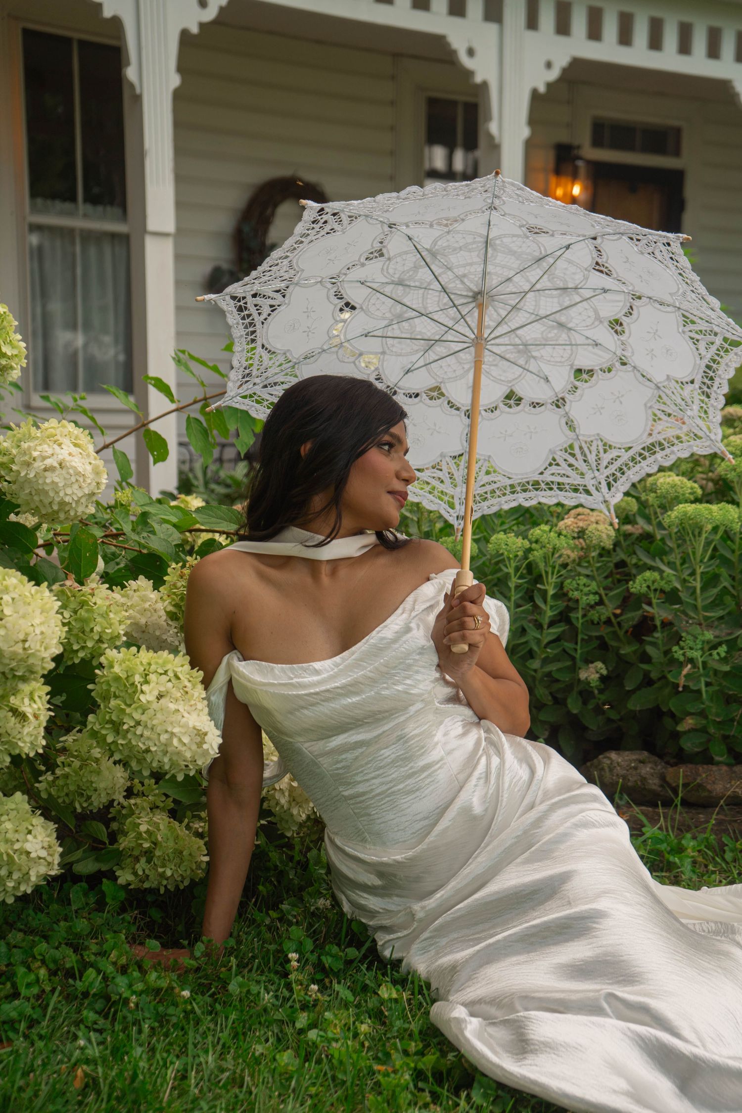 Bride in white off-shoulder gown sits among hydrangeas holding a delicate lace parasol outside a cottage.