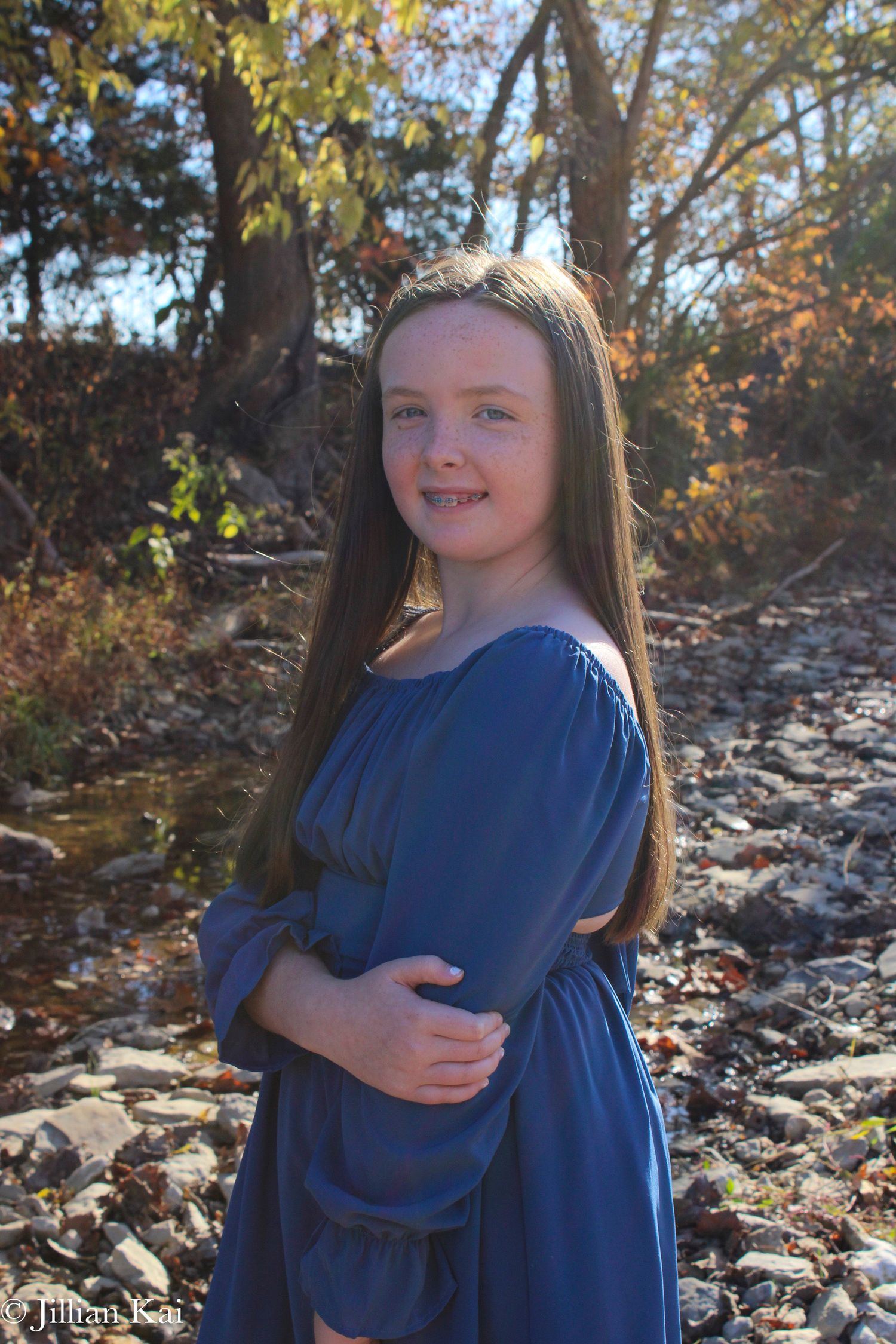 Young girl in blue dress smiling by rocky creek in autumn sunlight.