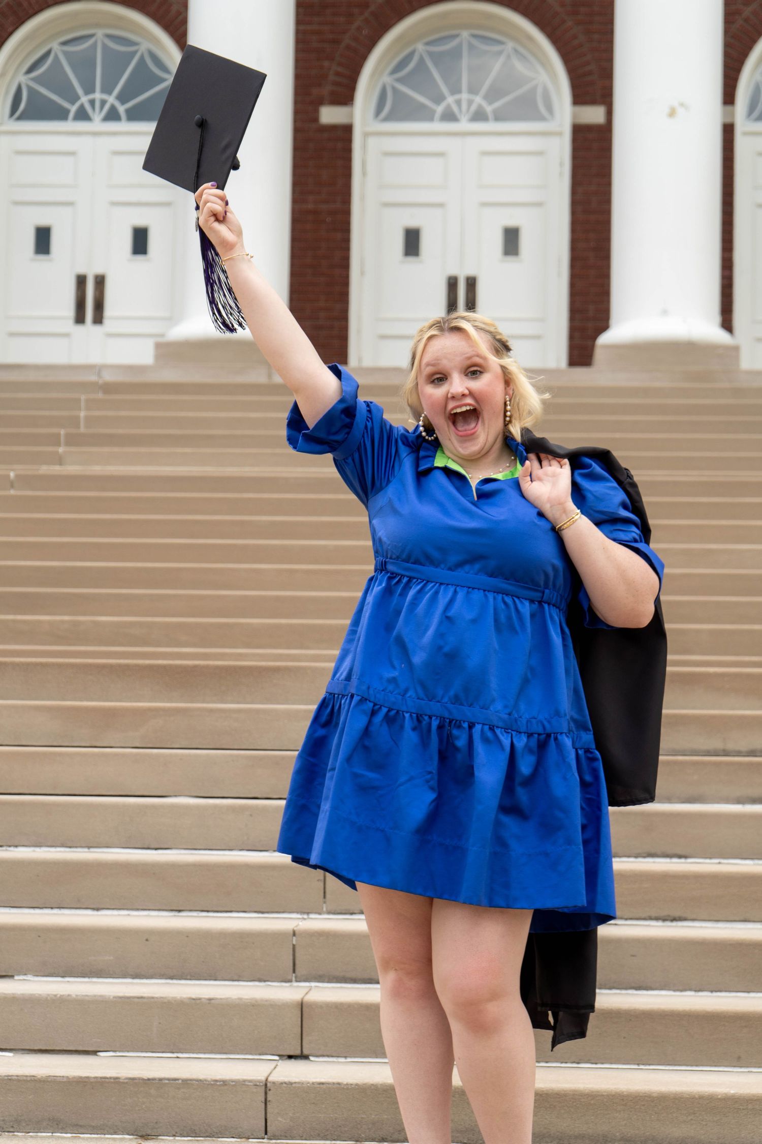 Excited blonde graduate in blue dress raising cap on university steps.