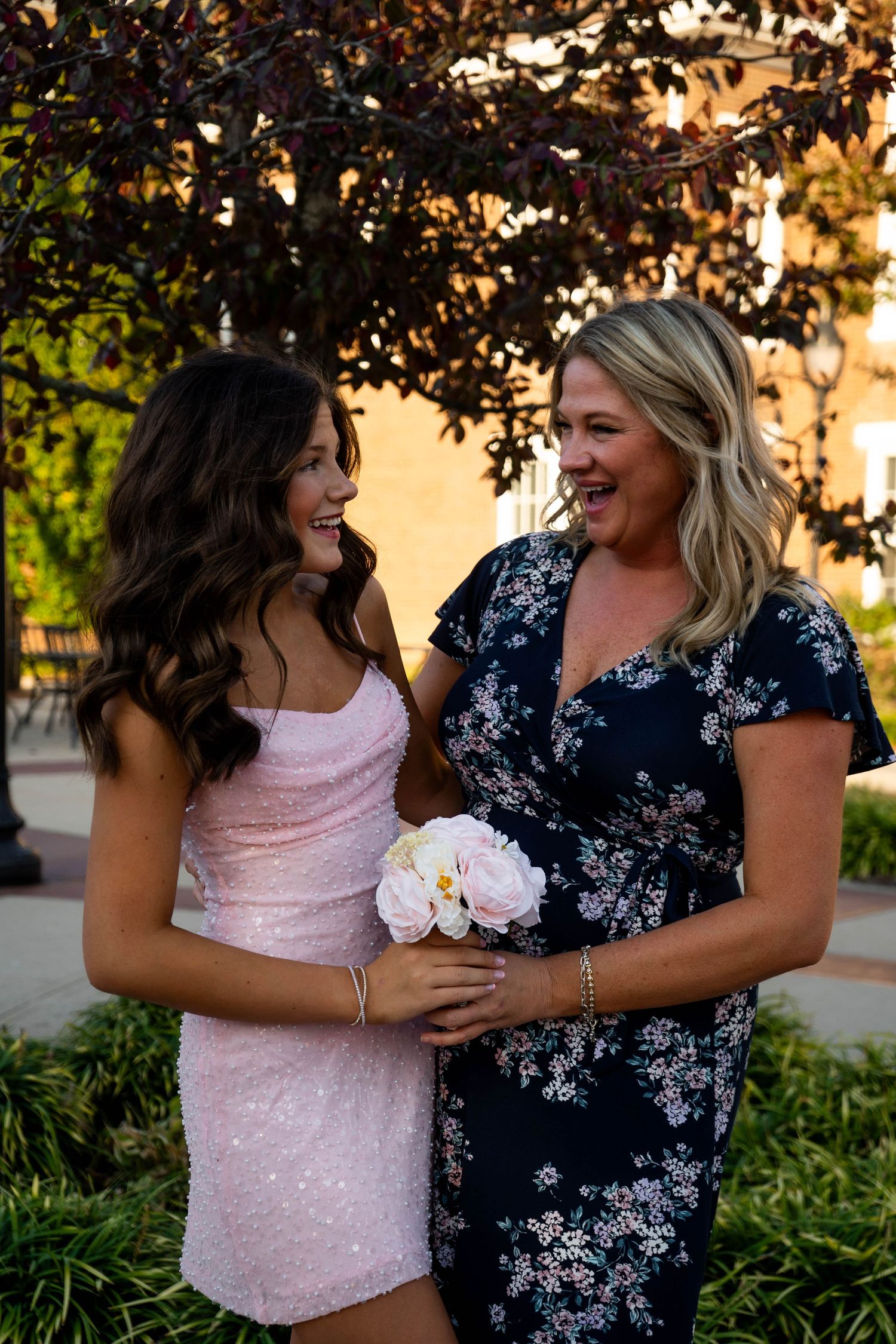 A teenage girl in a pink sequin dress and her smiling mother in a floral dress pose together outdoors.
