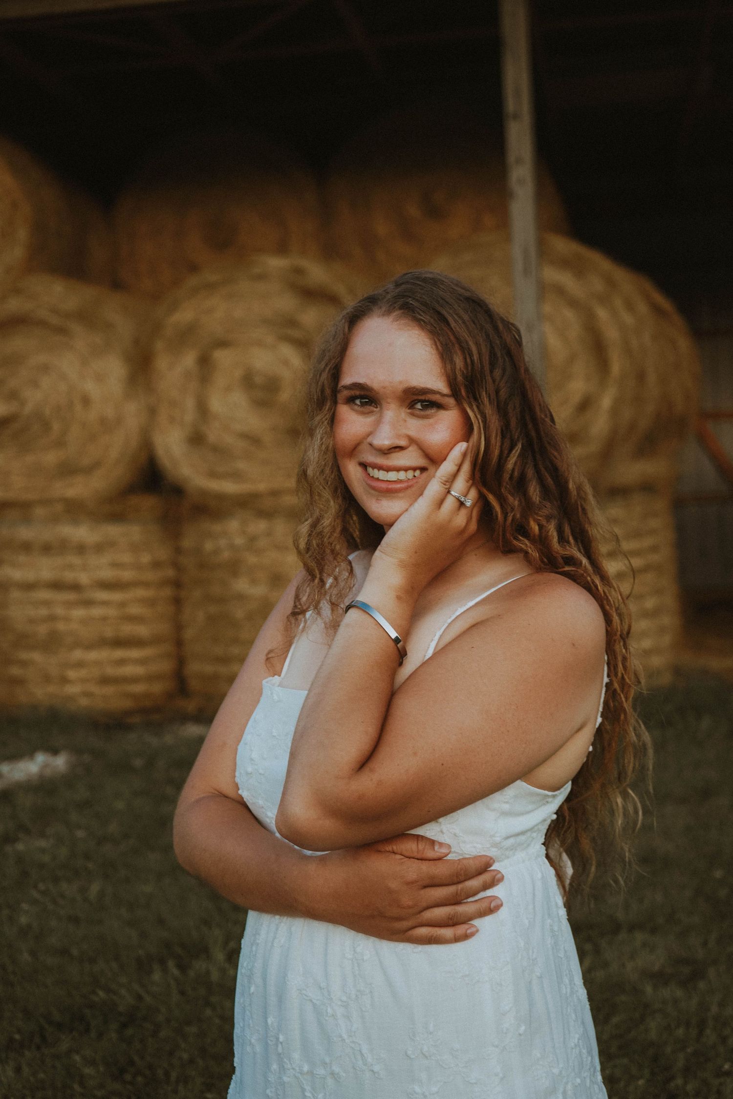 Happy young woman in white dress with hand on cheek posing by round hay bales.