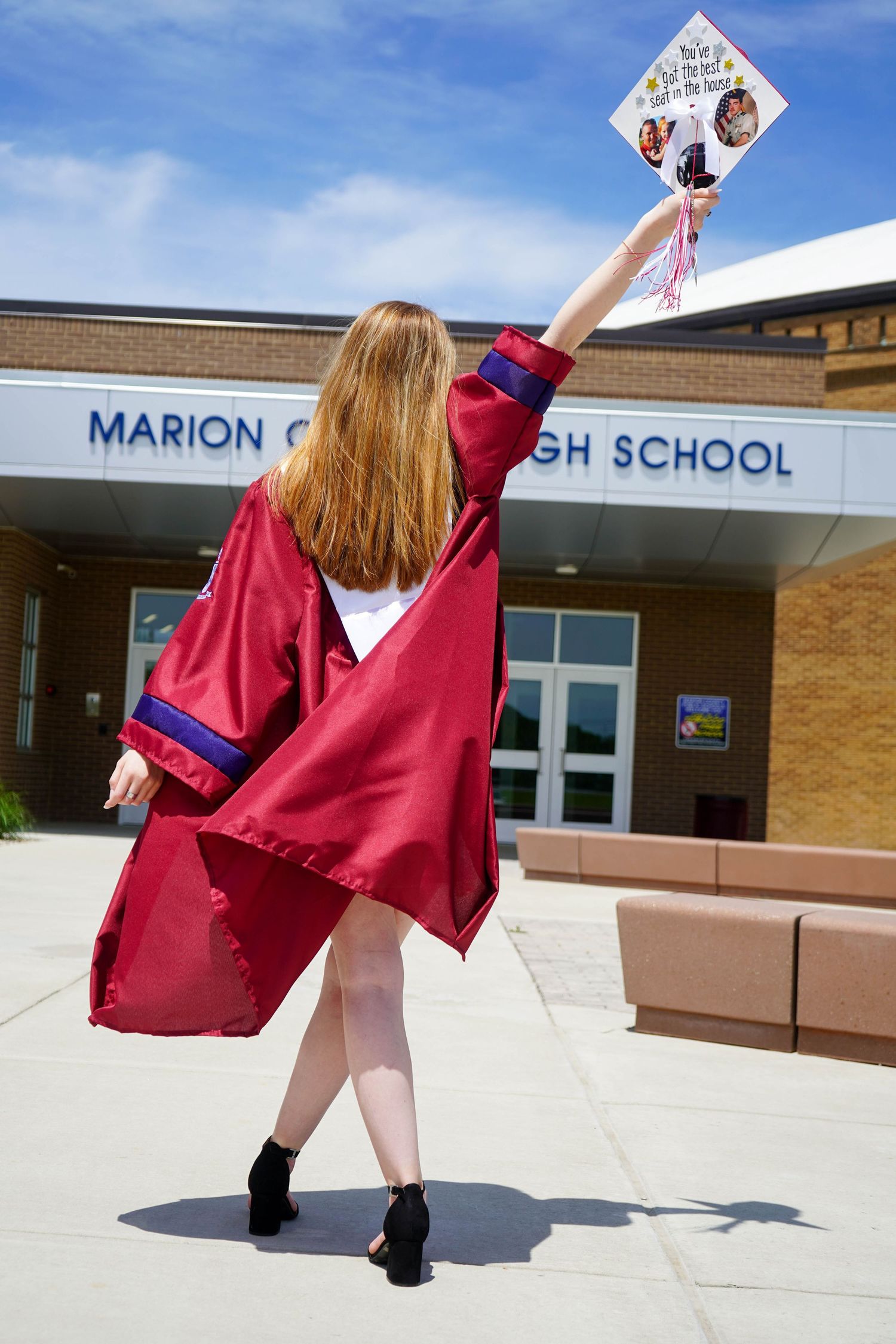 Female graduate in red cap and gown raising decorated mortarboard outside Marion County High School.