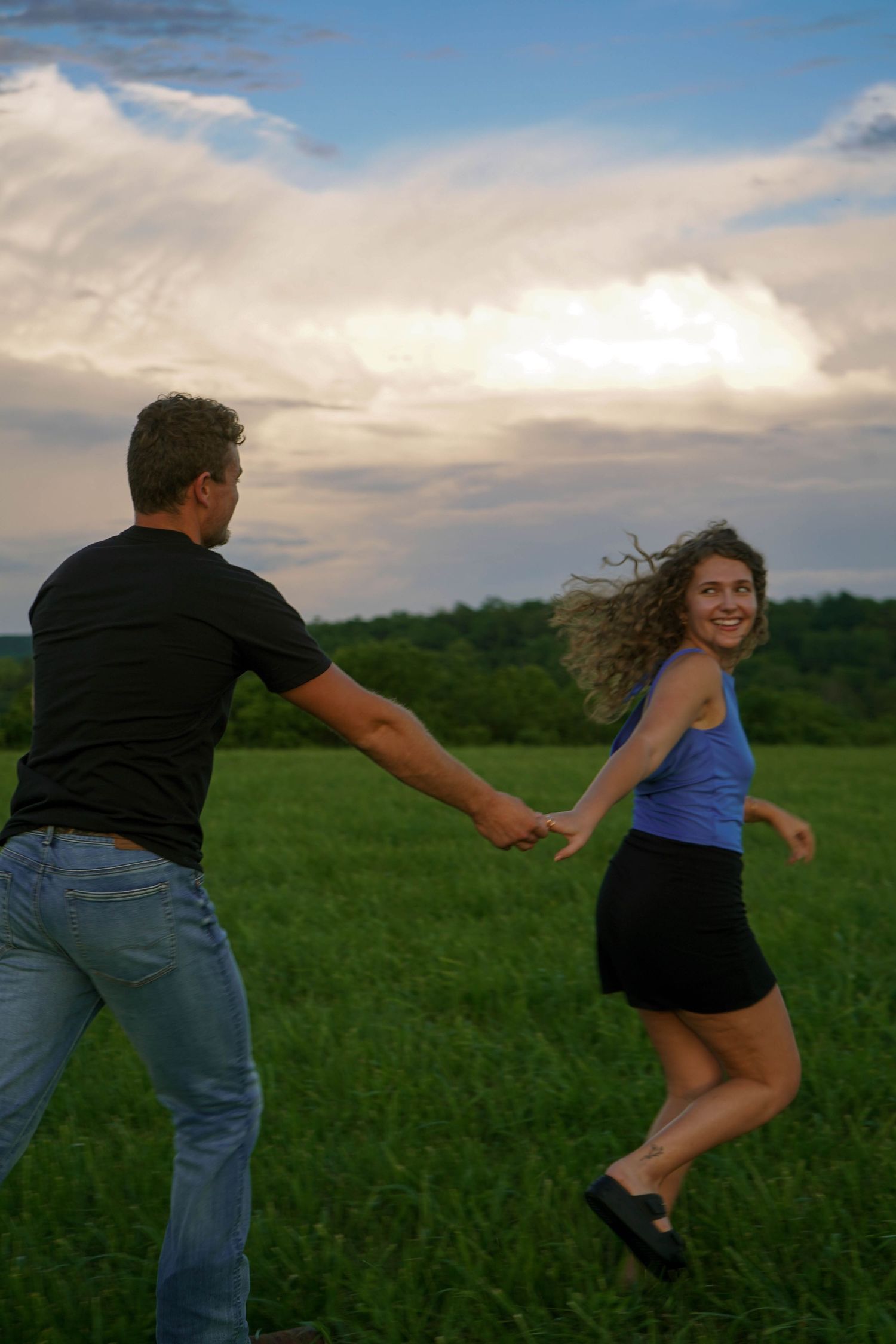 Joyful woman in blue top running through green field holding mans hand at sunset.