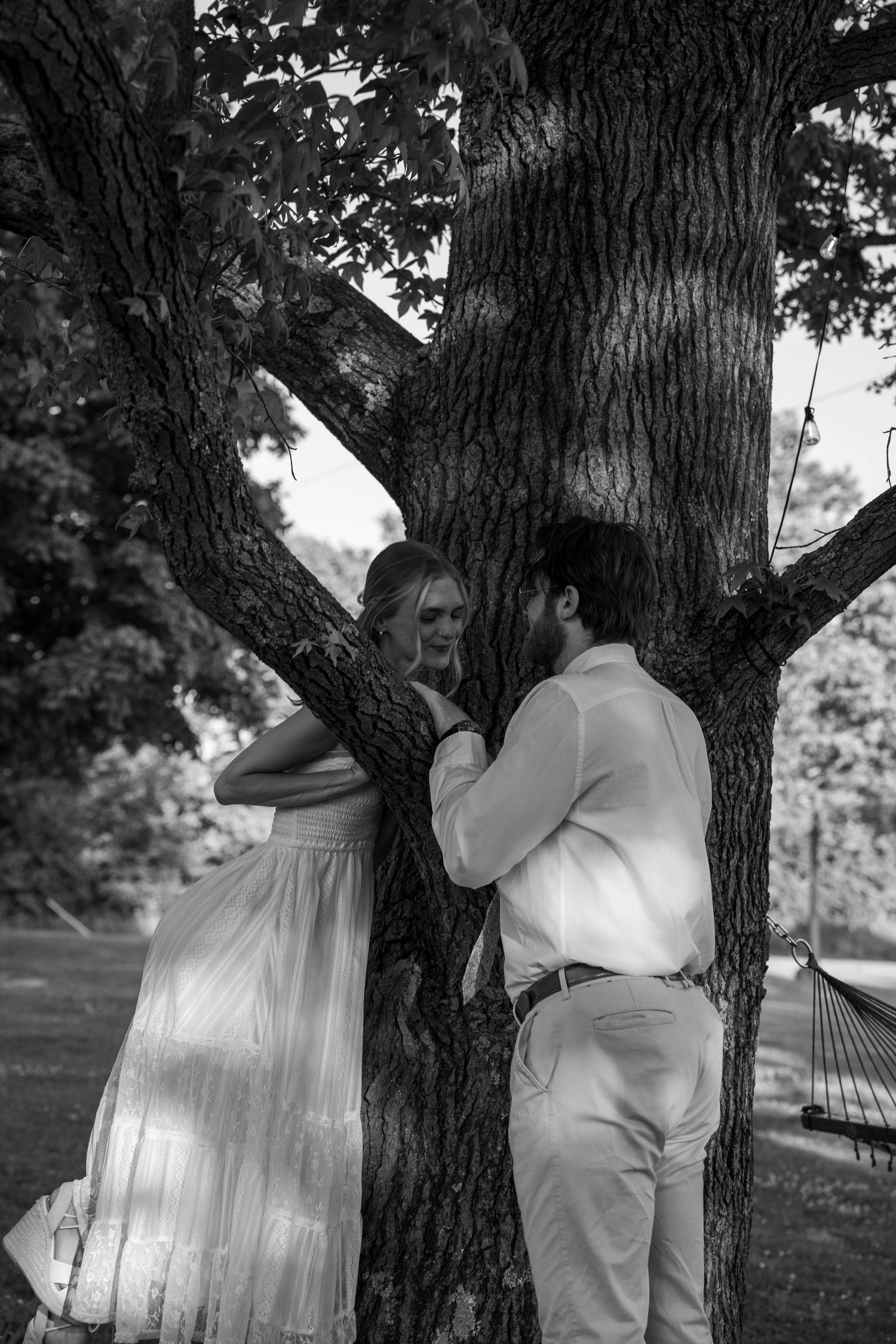 Black and white photo of couple sharing an intimate moment leaning against a large tree outdoors.