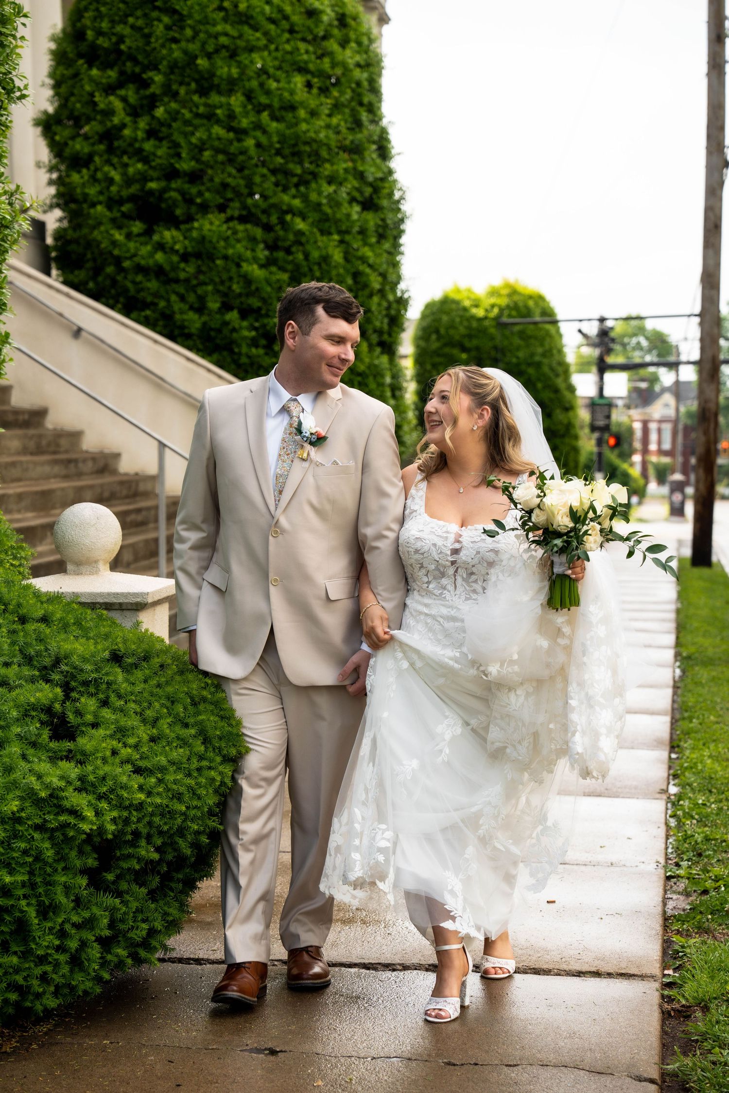 Bride and groom walk hand in hand on wet sidewalk, laughing, with lush green hedges behind them.
