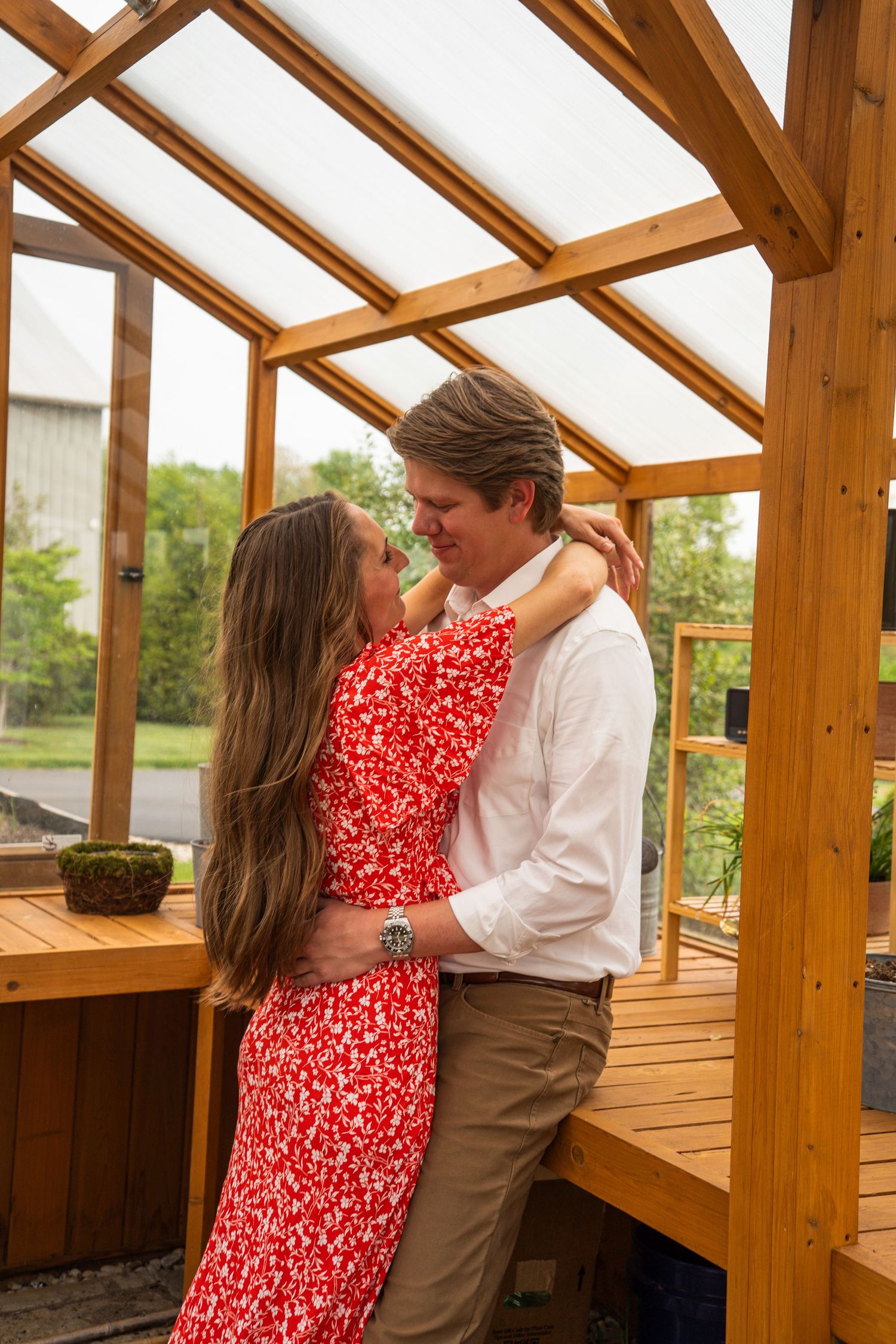 Happy engaged couple embracing and smiling inside a wooden greenhouse structure on a cloudy day.