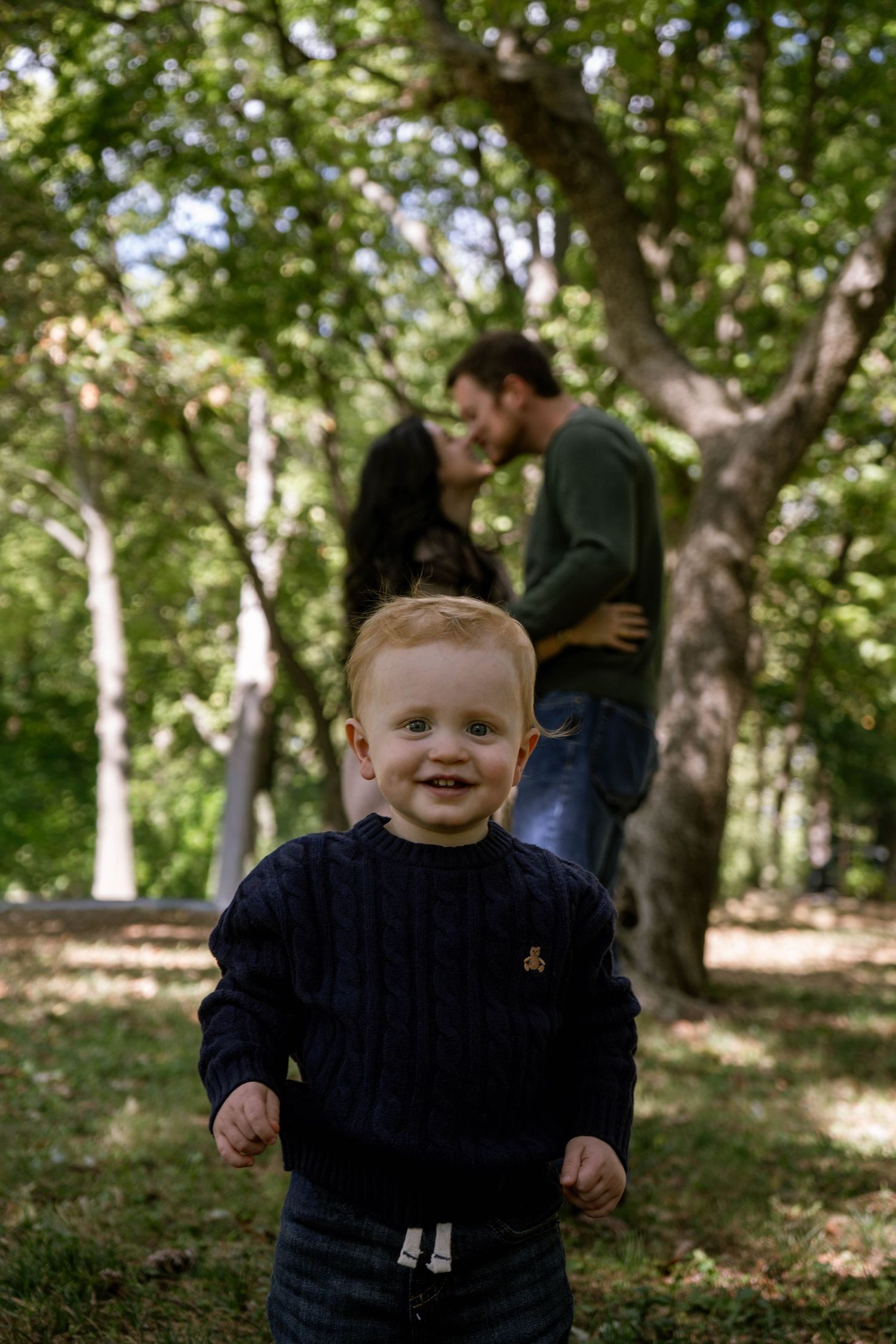 Smiling red-haired toddler in navy sweater stands in park while parents embrace under green trees behind him.