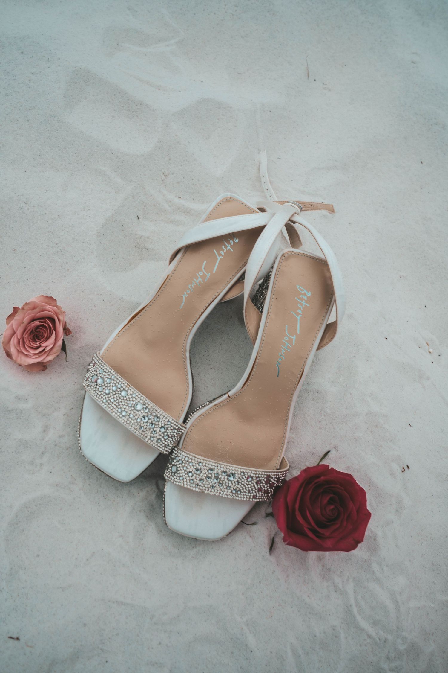 Bridal heels with crystal embellishments tied with ribbon surrounded by pink and red roses on sand.