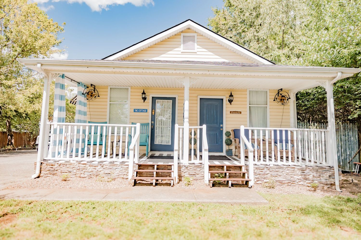 Charming yellow cottage with white wraparound porch, blue front door, stone accents, and lush trees in the background.