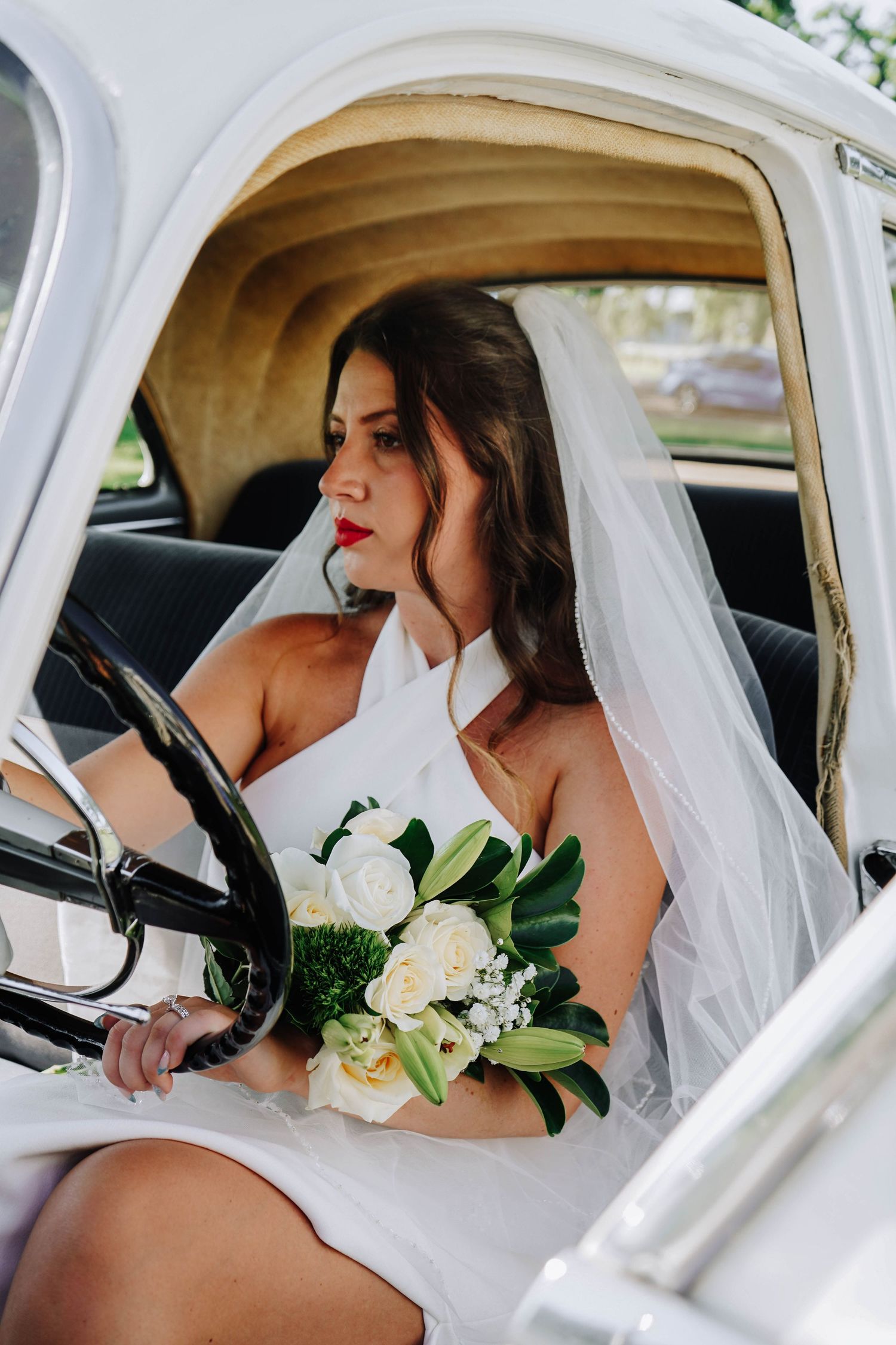 Bride in white halter wedding dress and veil sits in vintage white car holding bouquet of white roses and lilies.