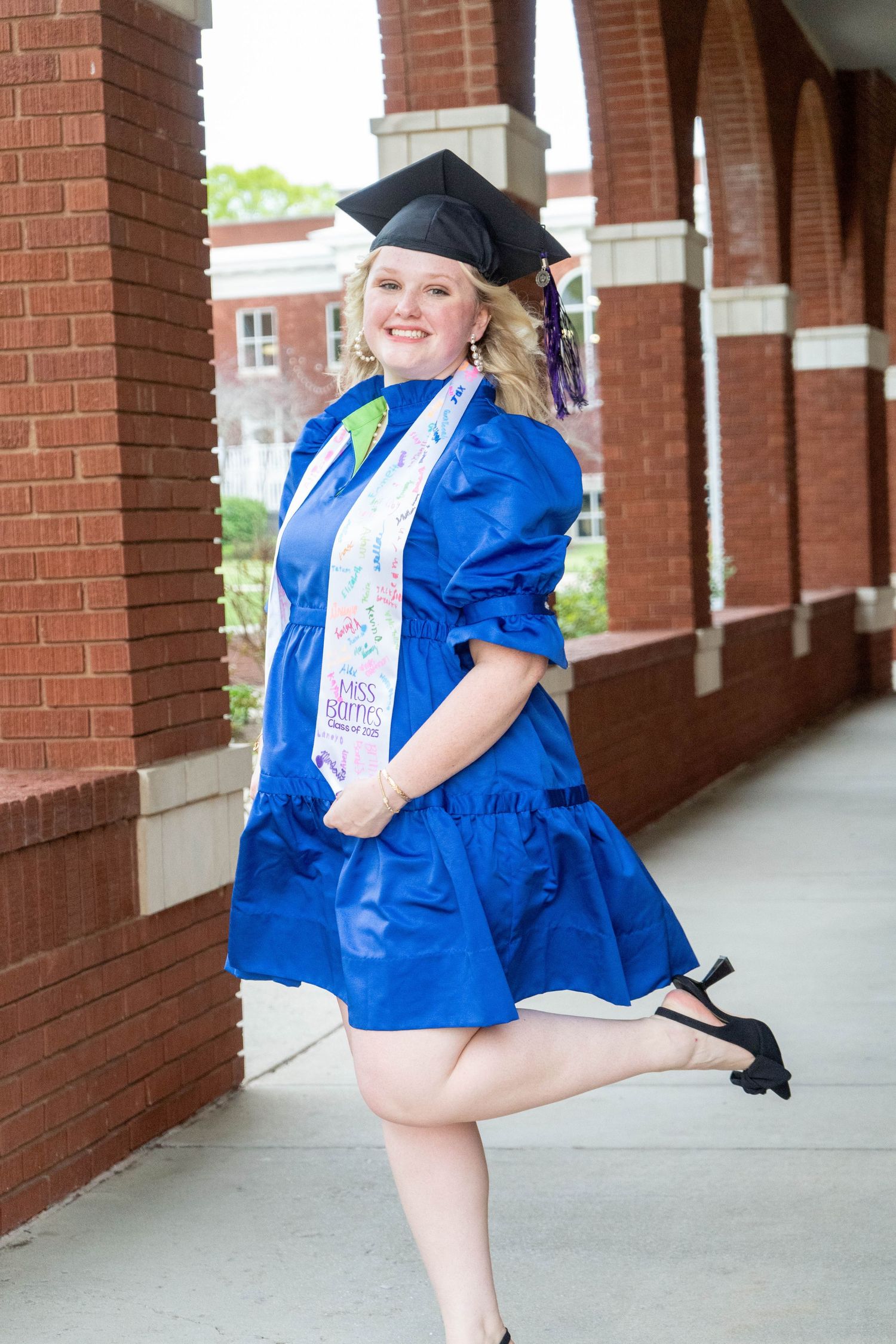 Joyful graduate in blue dress and cap posing in brick archway corridor.