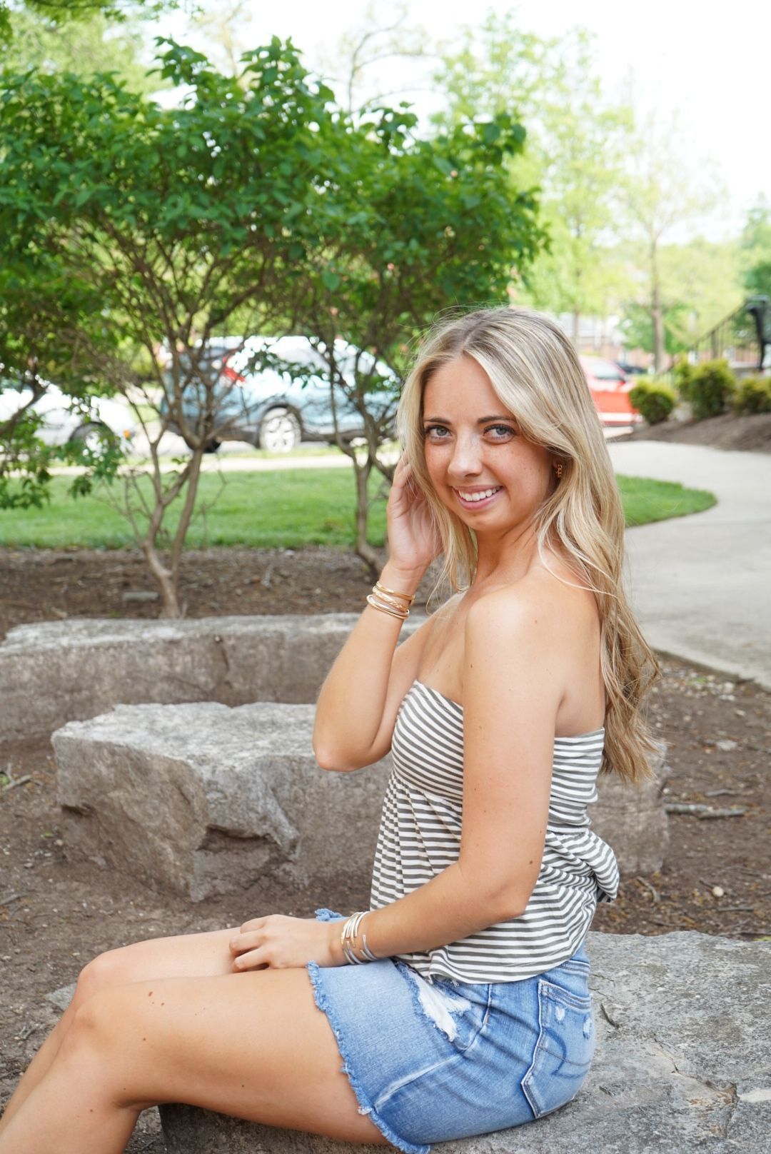 Smiling blonde woman in striped top and denim shorts sitting on stone steps outdoors.