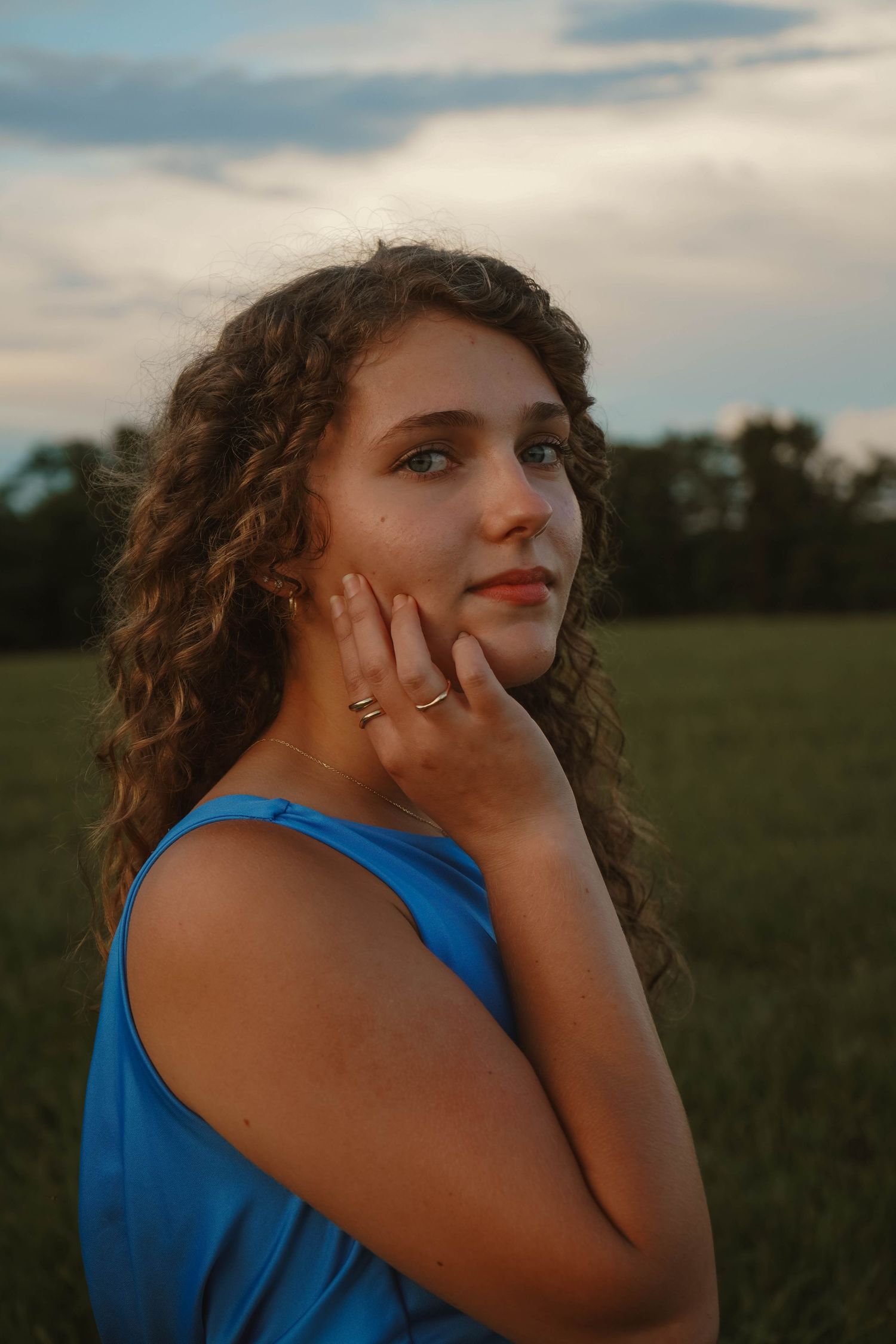 Young woman with curly hair in blue dress poses thoughtfully in golden hour field.