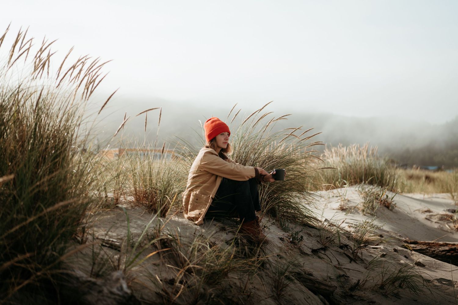 Person in red beanie sitting on sandy dunes surrounded by tall grass in a misty coastal landscape.