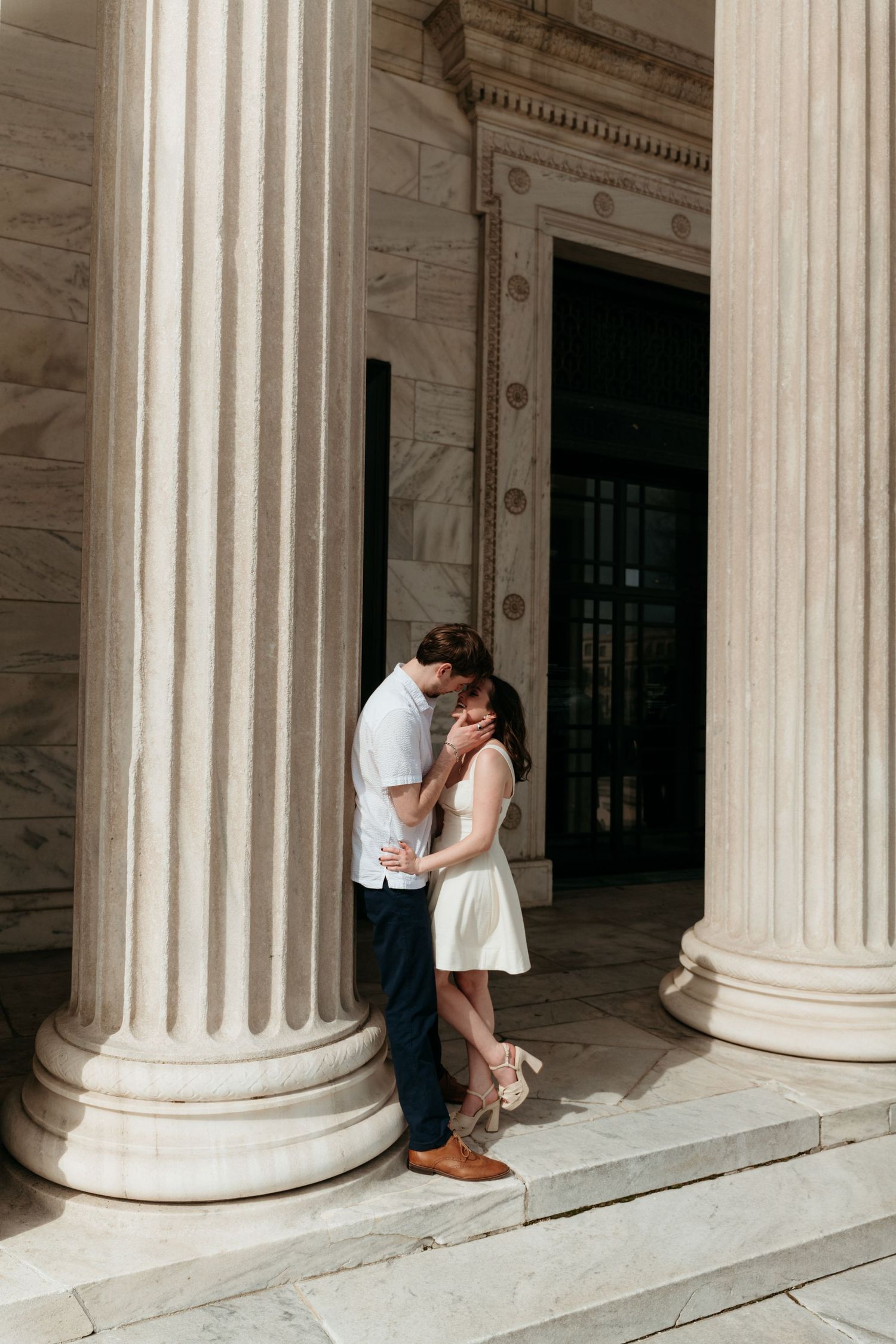 Couple sharing a romantic kiss between grand marble columns at a neoclassical building entrance, woman in white dress.