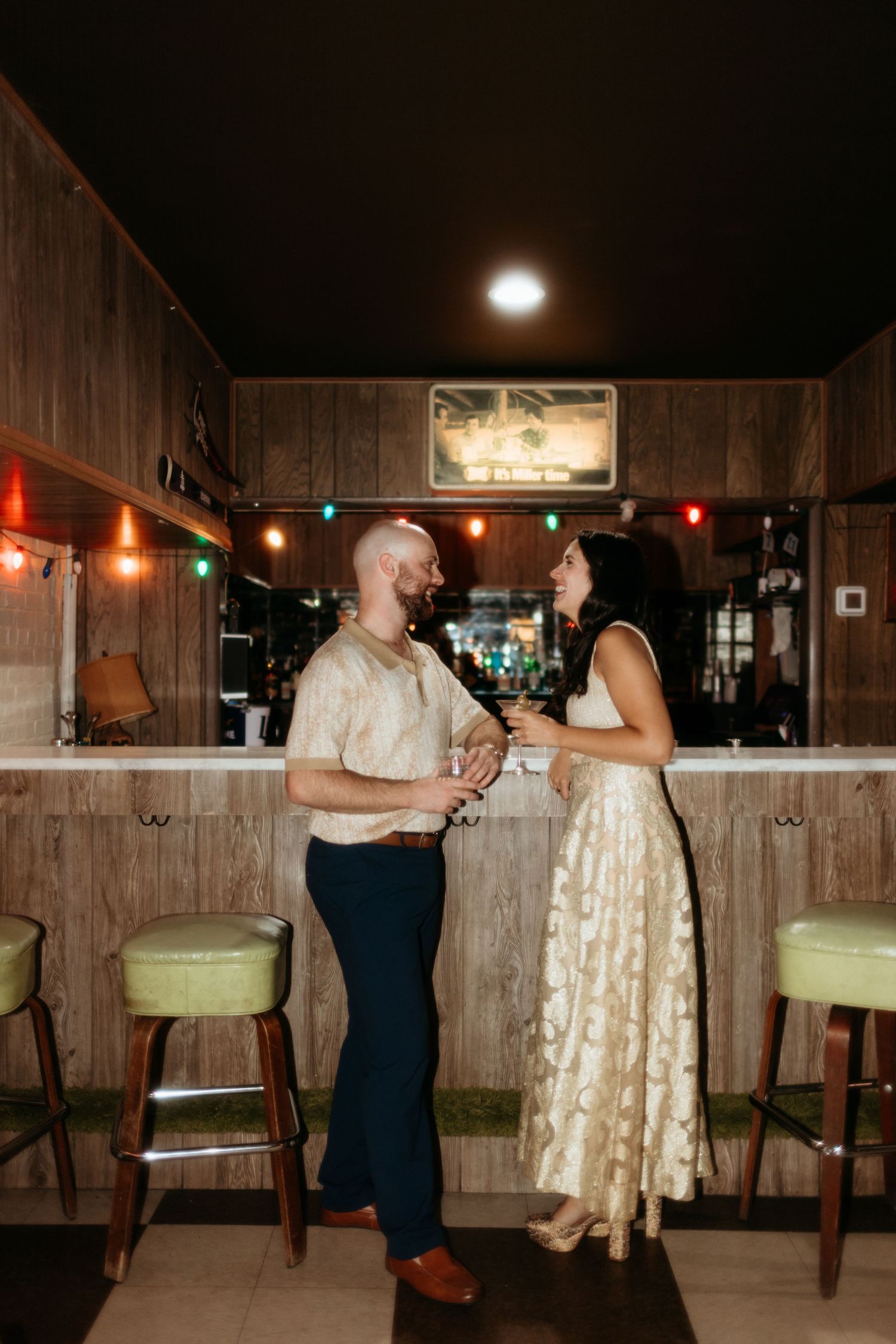 Couple sharing drinks at a vintage wood-paneled bar with green stools and string lights.