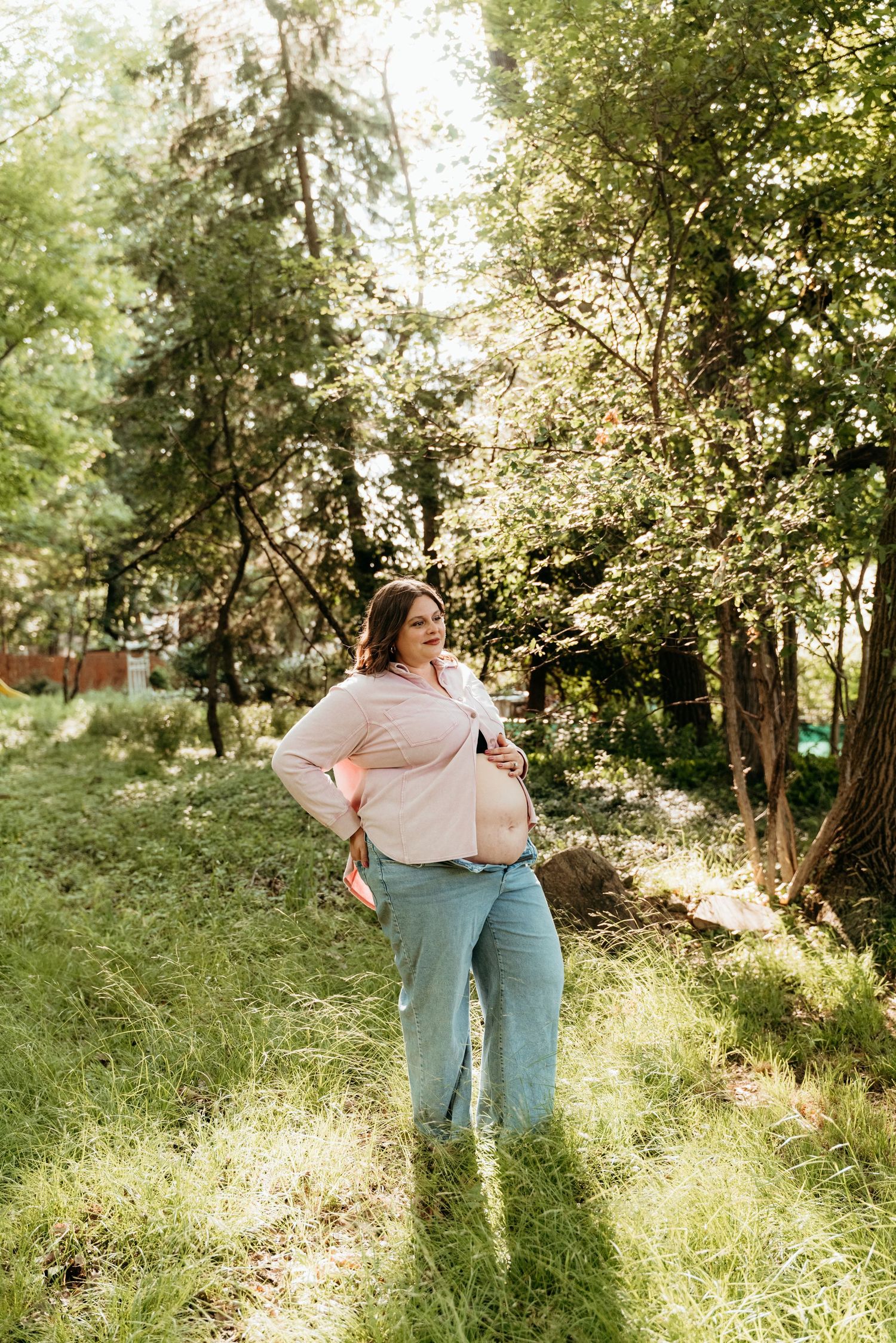 Pregnant woman in pink top and jeans standing outdoors in a sunlit green forest setting.