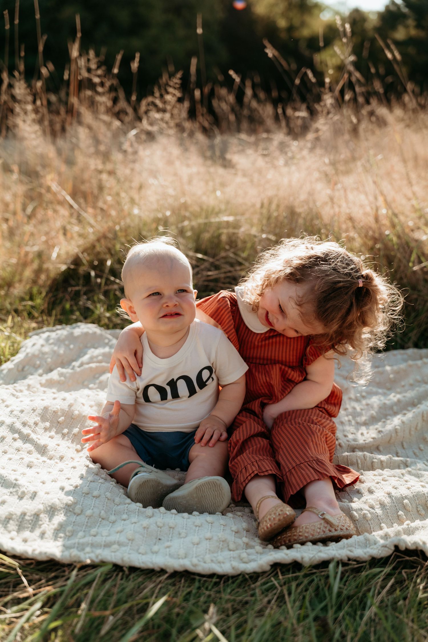 Two young siblings sit together on a white blanket in a sunny field, older girl in red hugging baby wearing \