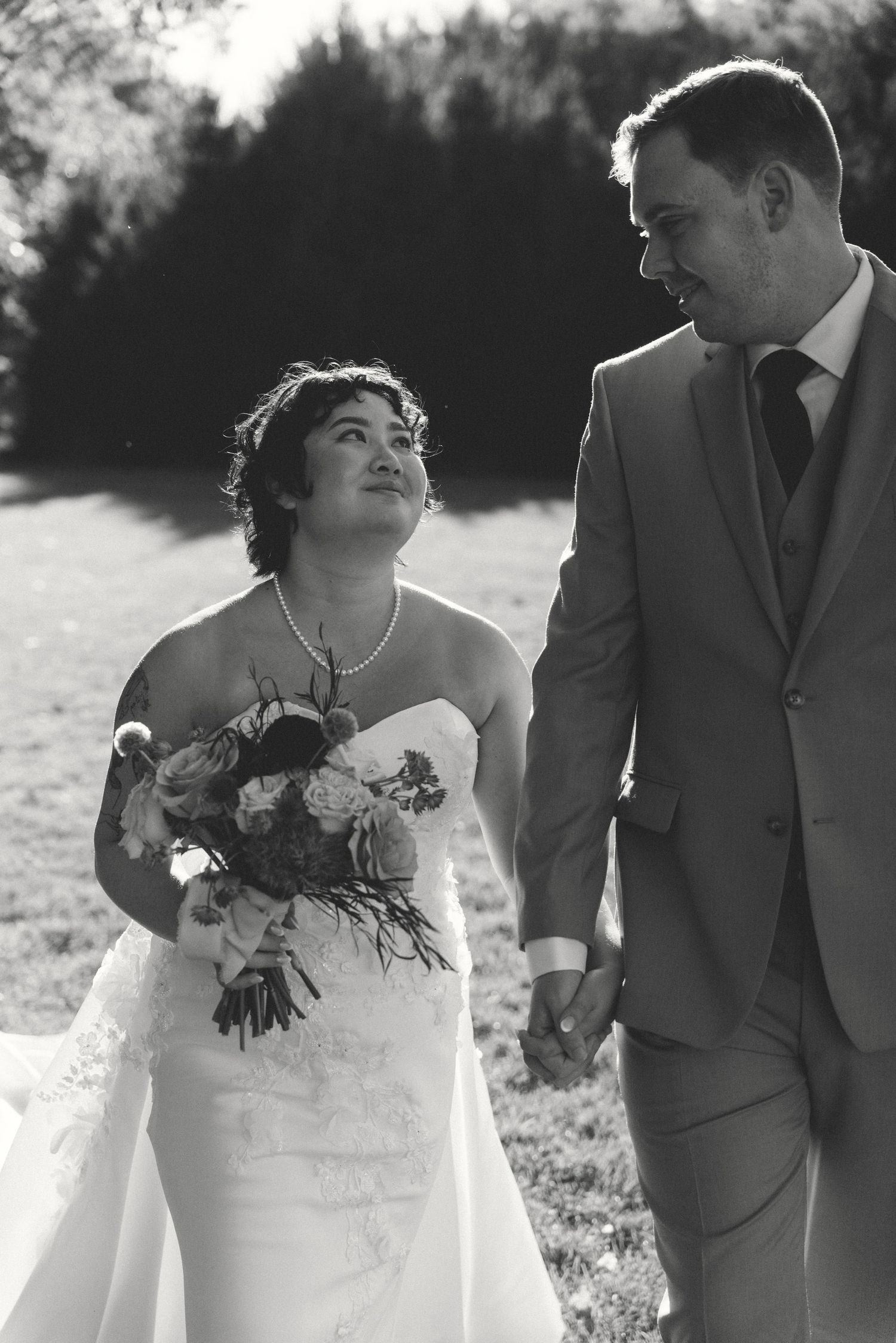 Black and white photo of a smiling bride holding a bouquet and groom in a suit walking hand in hand outdoors.