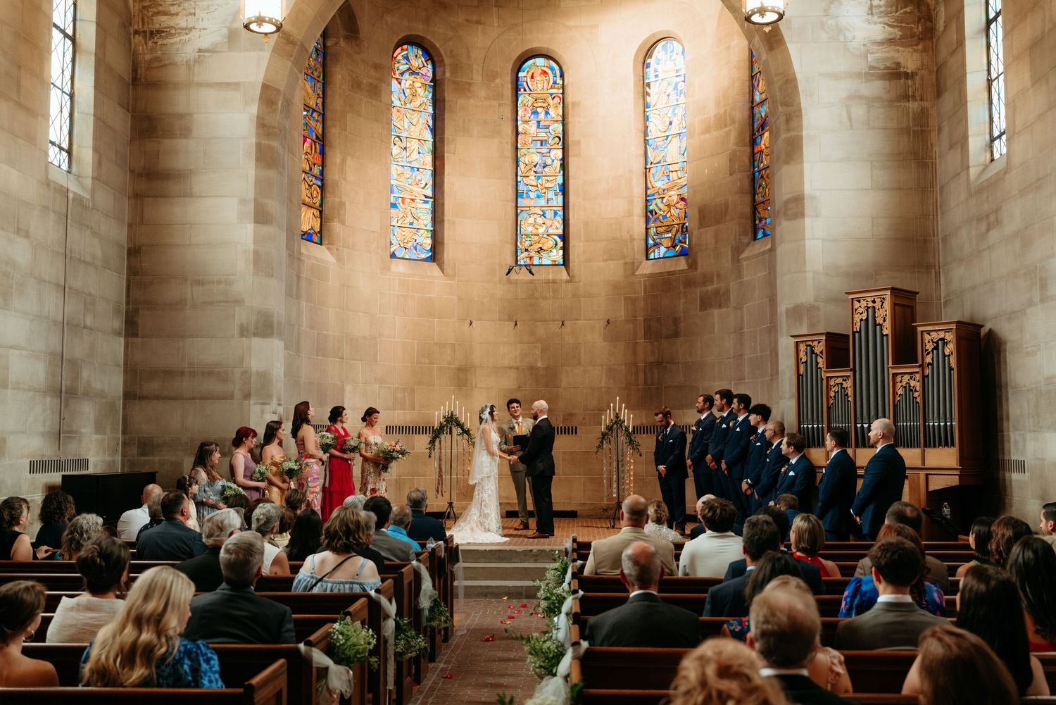 Elegant church wedding ceremony with stained glass windows, wooden pews, and wedding party at the altar.
