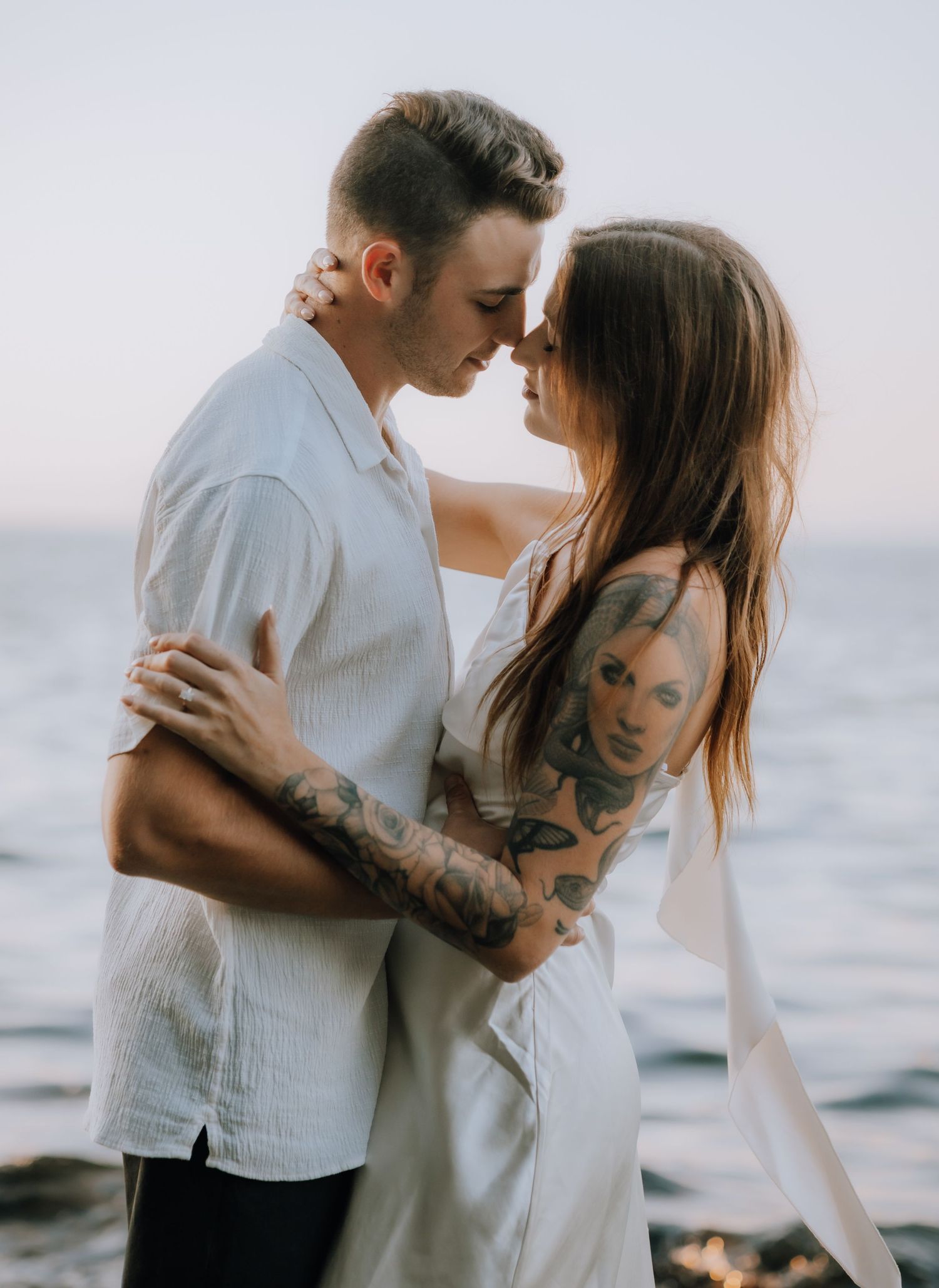 A romantic couple embraces on a misty beach at sunset wearing white and neutral tones.