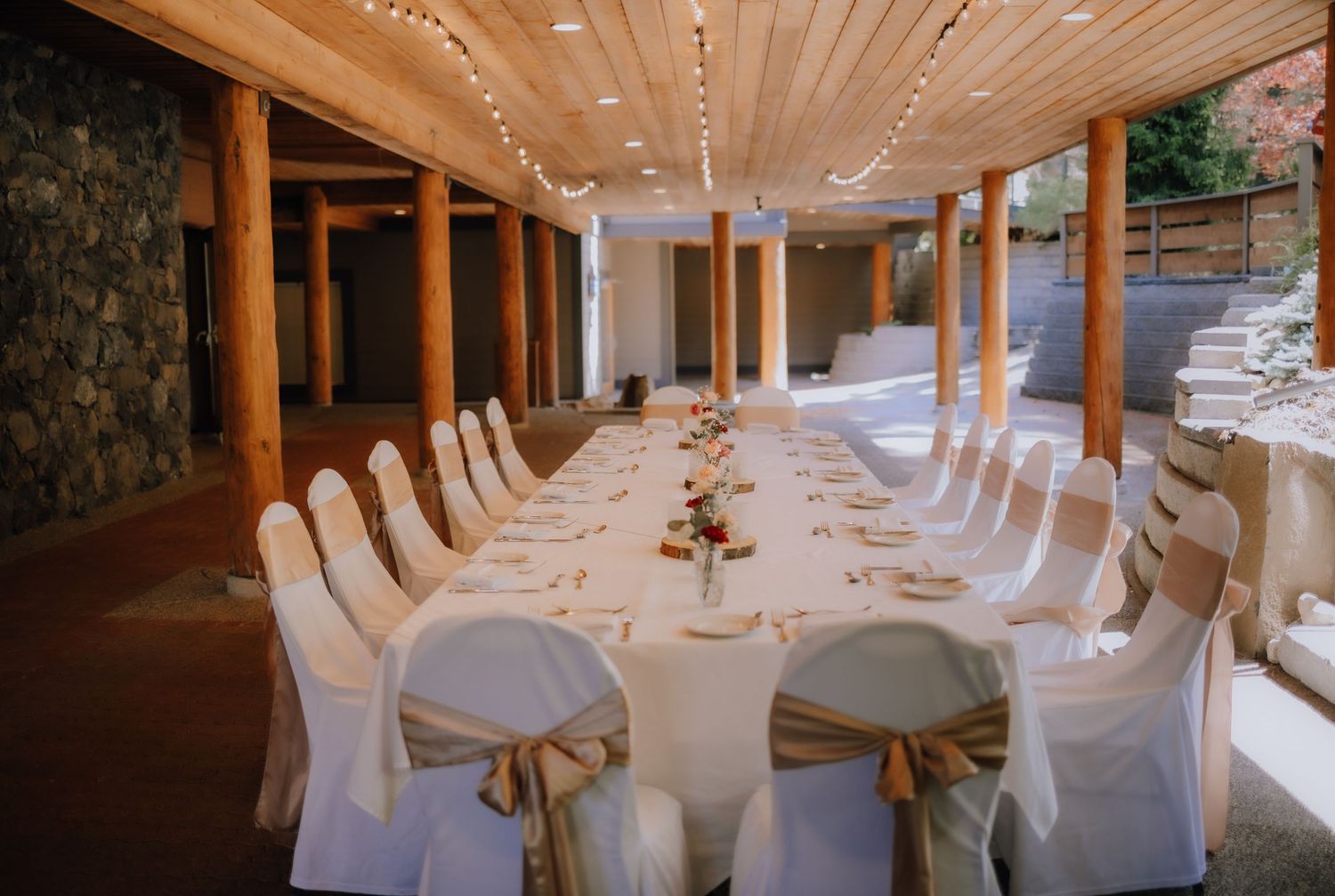 Elegant wedding reception table setup with white chair covers and gold sashes in a wooden beam venue with stone walls.
