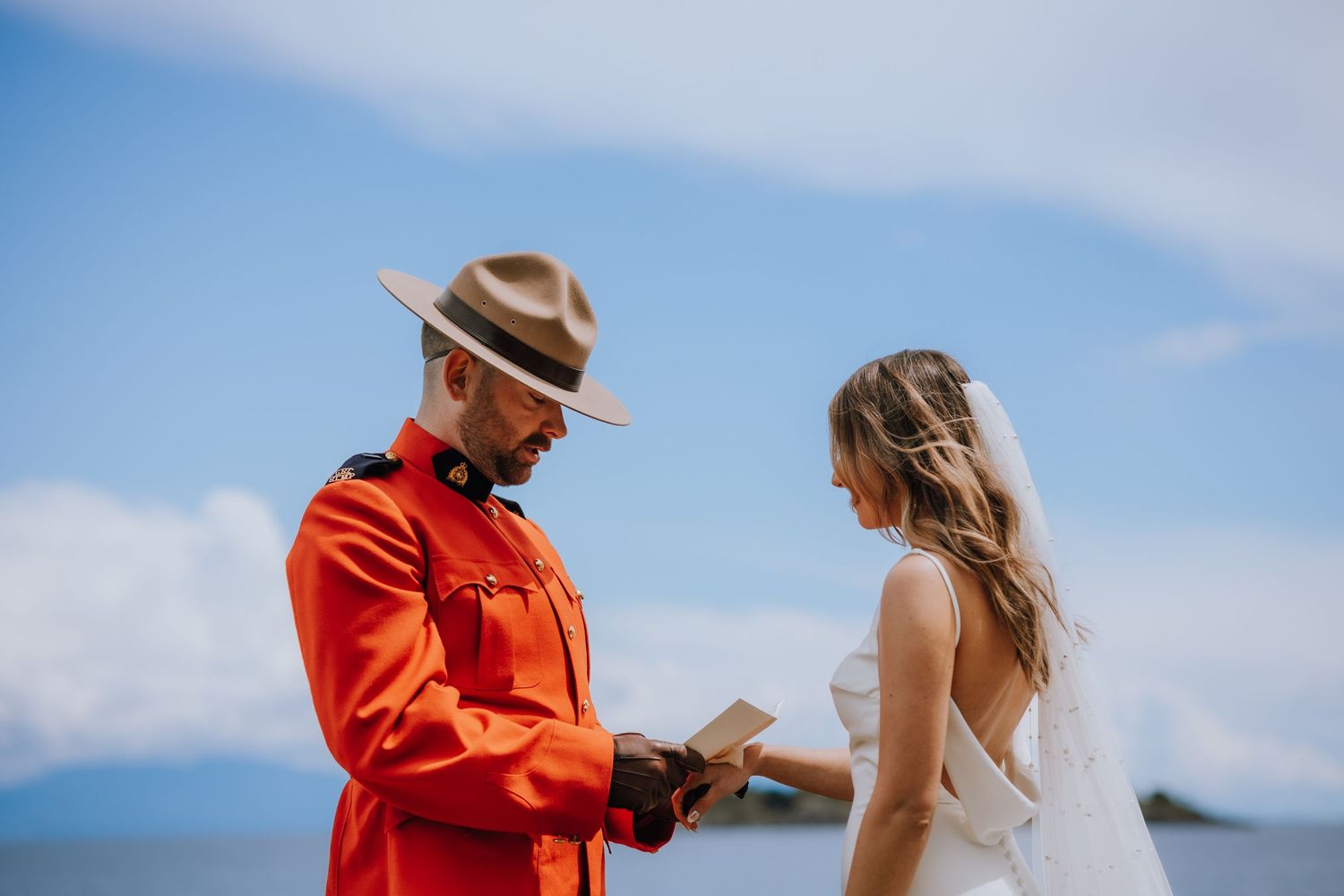Two people share a moment during a wedding ceremony against a scenic mountain backdrop by a lake on a bright sunny day.