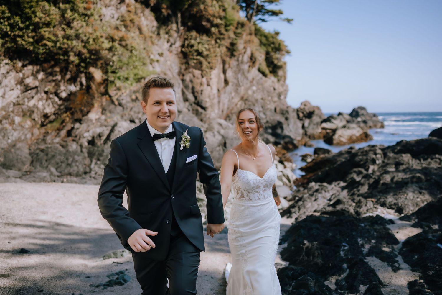 A couple in wedding attire walks along a scenic rocky beach coastline with ocean views and cliffs in the background.