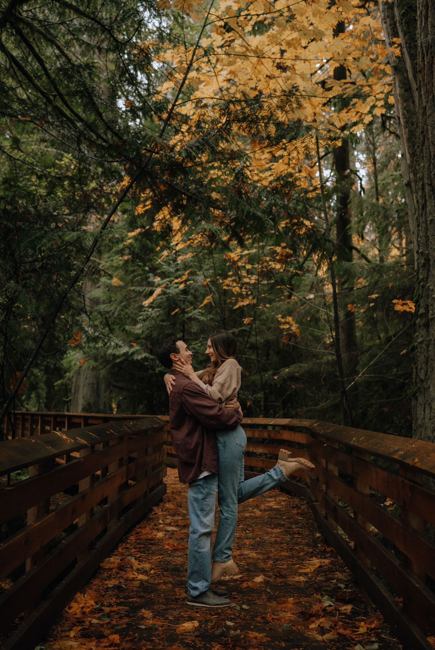 A couple shares a romantic moment on a wooden bridge surrounded by autumn foliage in a forest setting.