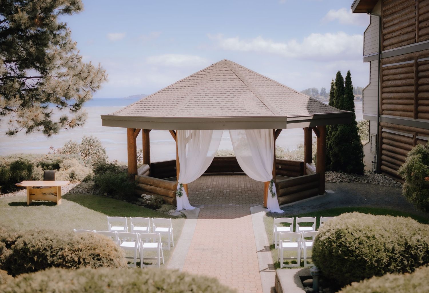 An outdoor wedding gazebo with white curtains and chairs set up on a stone path, surrounded by manicured gardens.