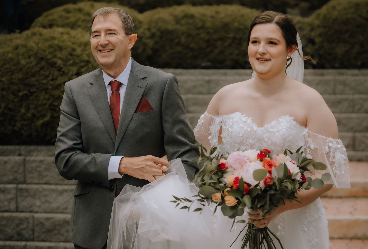 Wedding photo of two people standing on stone steps with greenery backdrop and bride holding colorful floral bouquet.