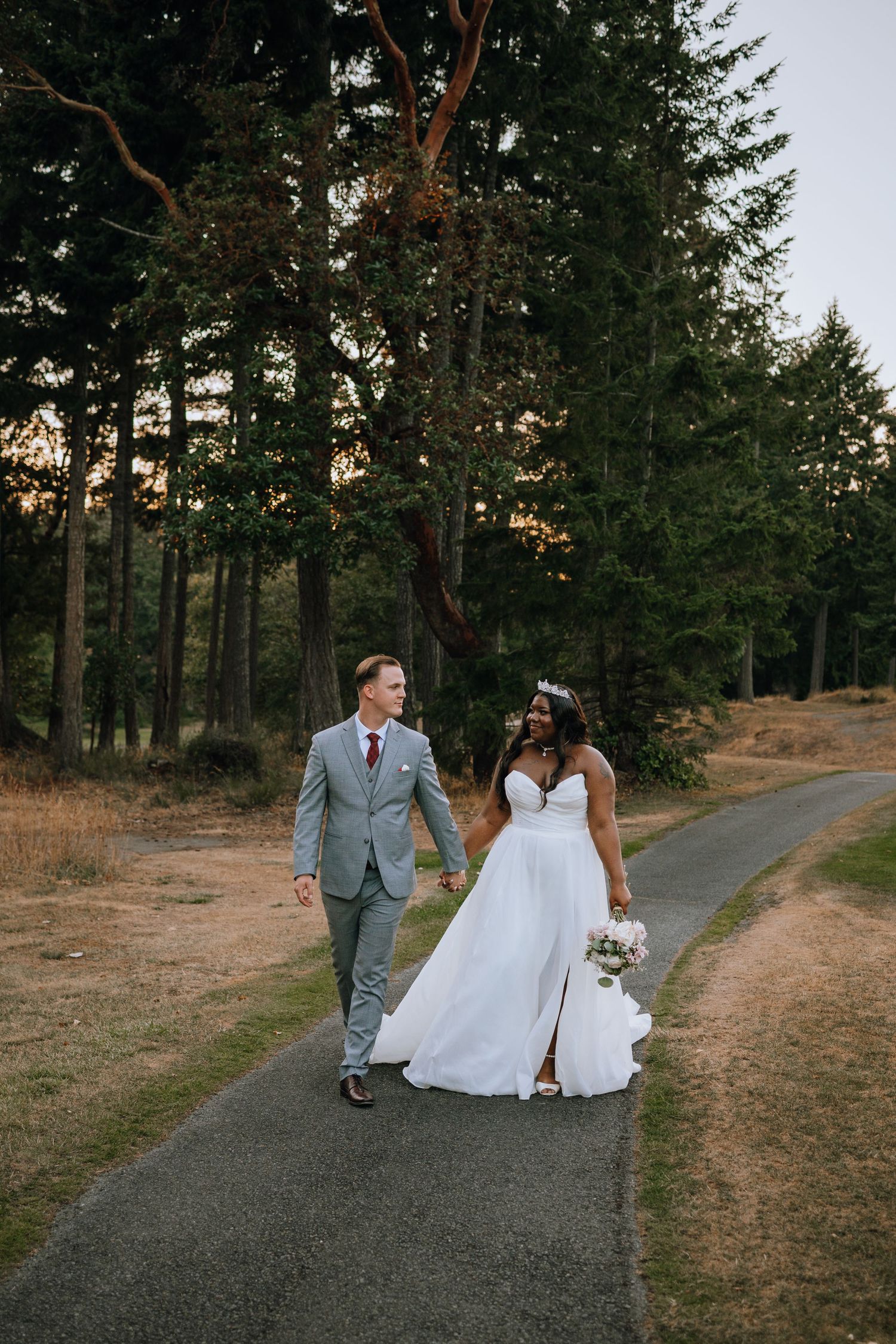 A couple walks hand in hand down a winding path surrounded by tall evergreen trees during a sunset wedding photo session.