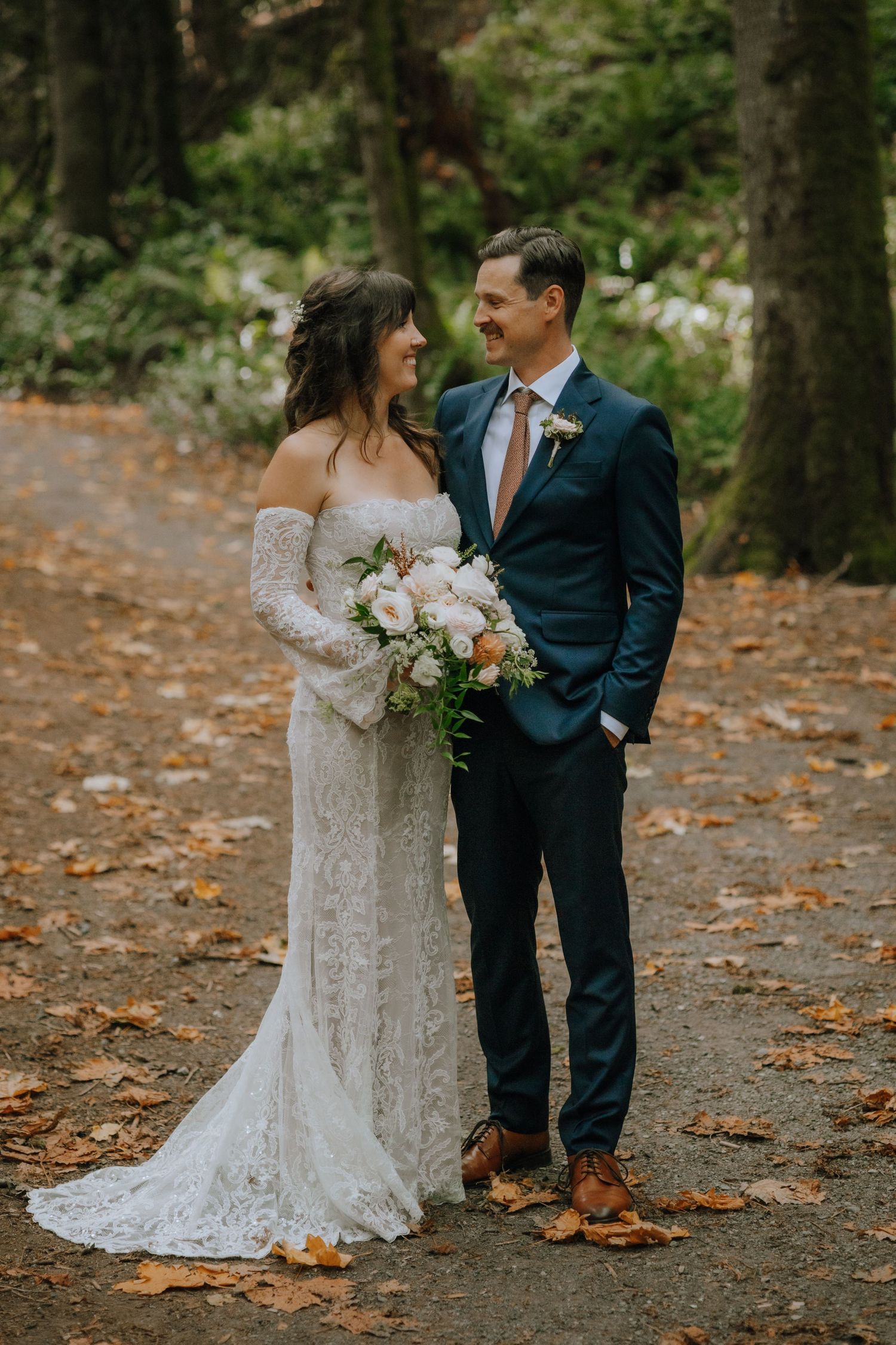 A couple shares a joyful moment on a forest path during their autumn wedding, surrounded by fallen leaves.