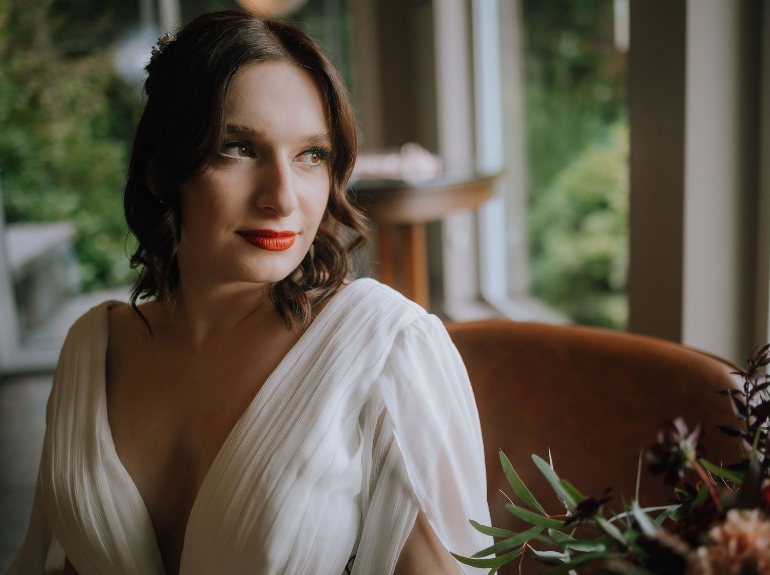A bride in a white wedding dress poses in natural light by a window during a wedding photoshoot.